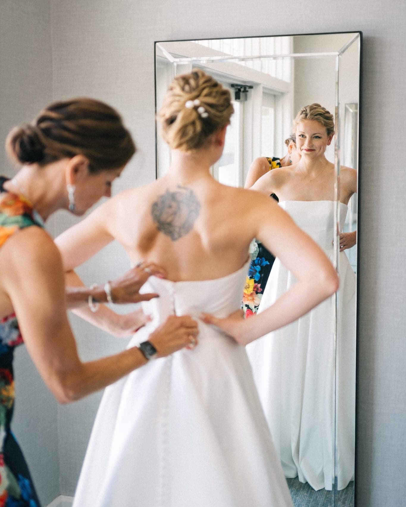 A bride in a strapless wedding dress is being assisted by a woman in a floral dress as they look at themselves in a mirror.