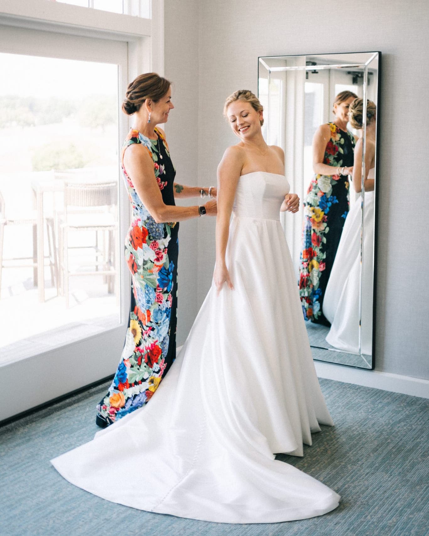 A woman in a floral dress helps another woman in a white strapless gown in a room with a large mirror and a window.