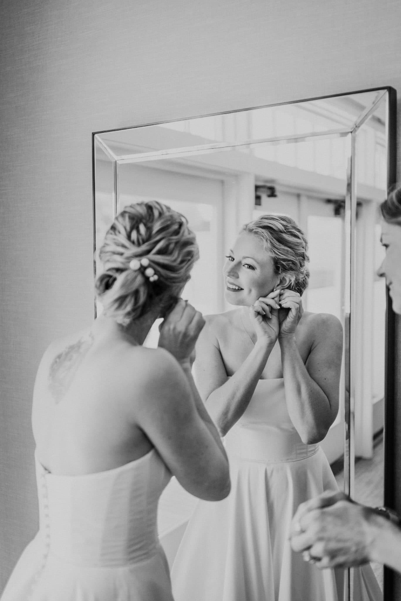 A bride adjusts her earrings in front of a mirror, her reflection showing her smiling face.