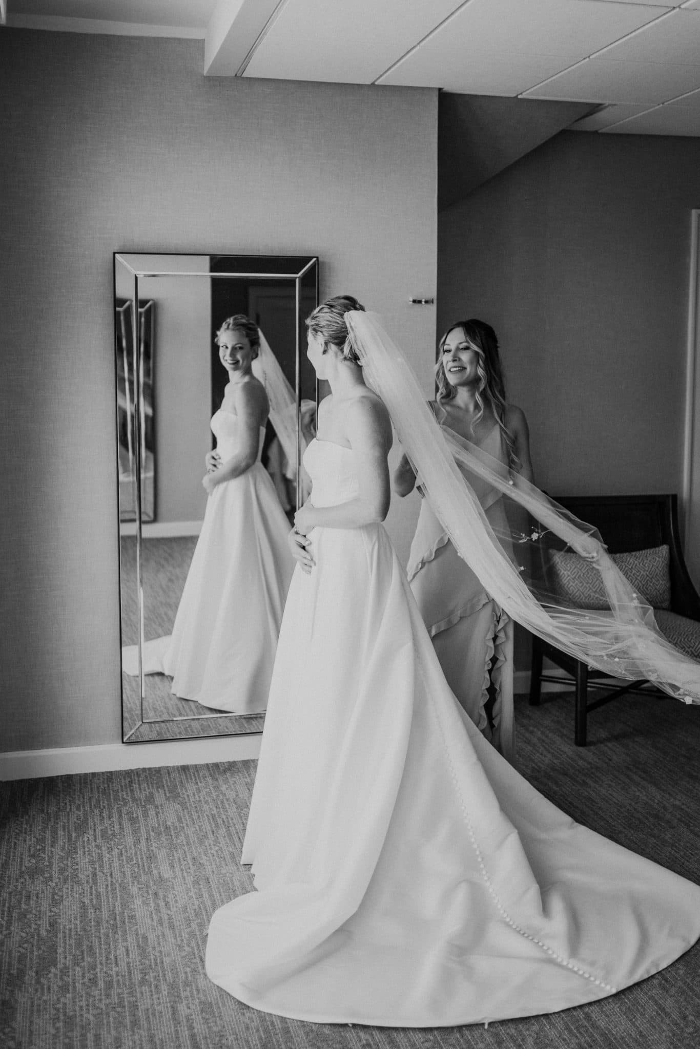 A bride in a white gown stands in front of a mirror, assisted by a woman adjusting her veil.