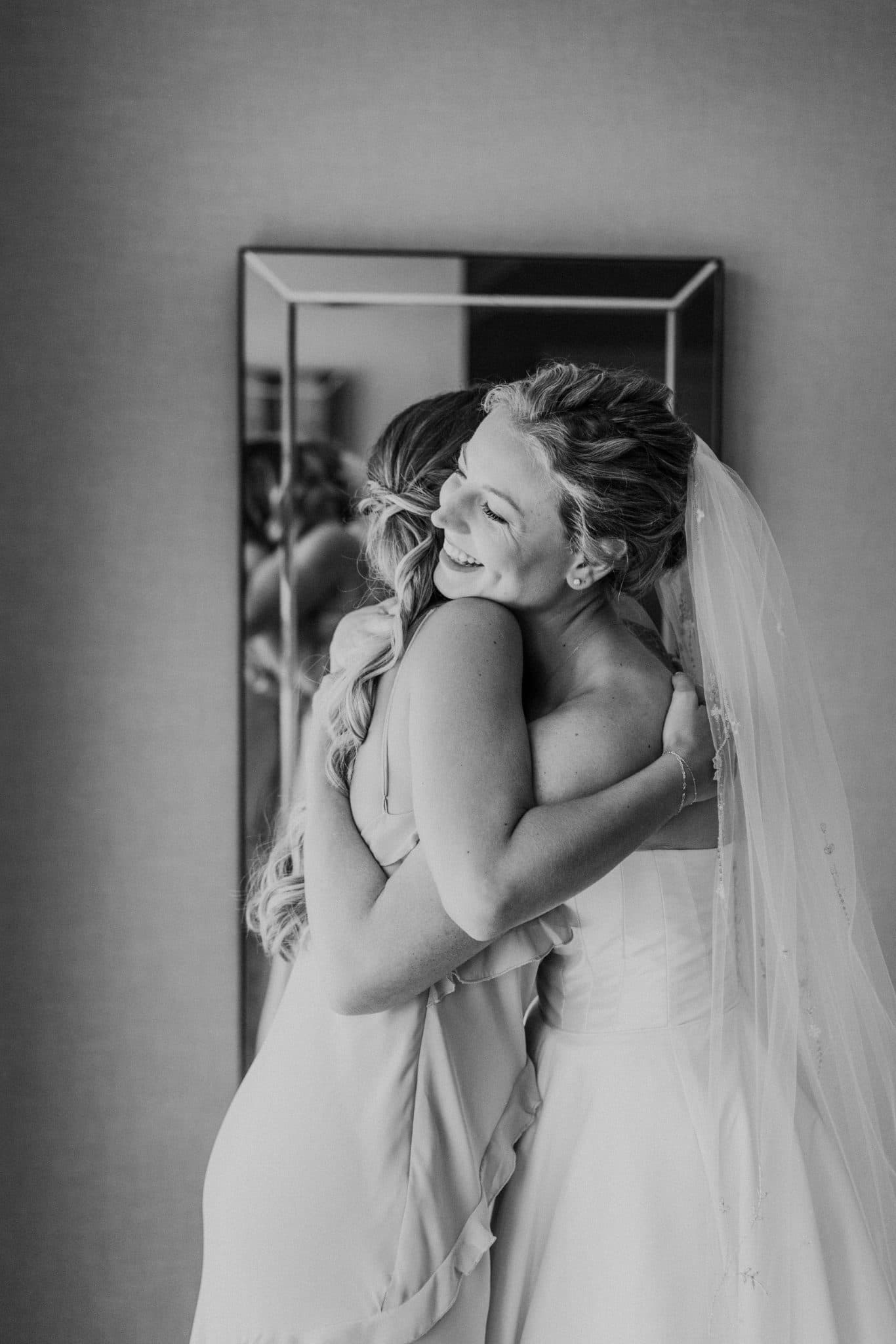 A bride in a wedding dress hugs a woman in a light-colored dress, both smiling, in front of a mirror.