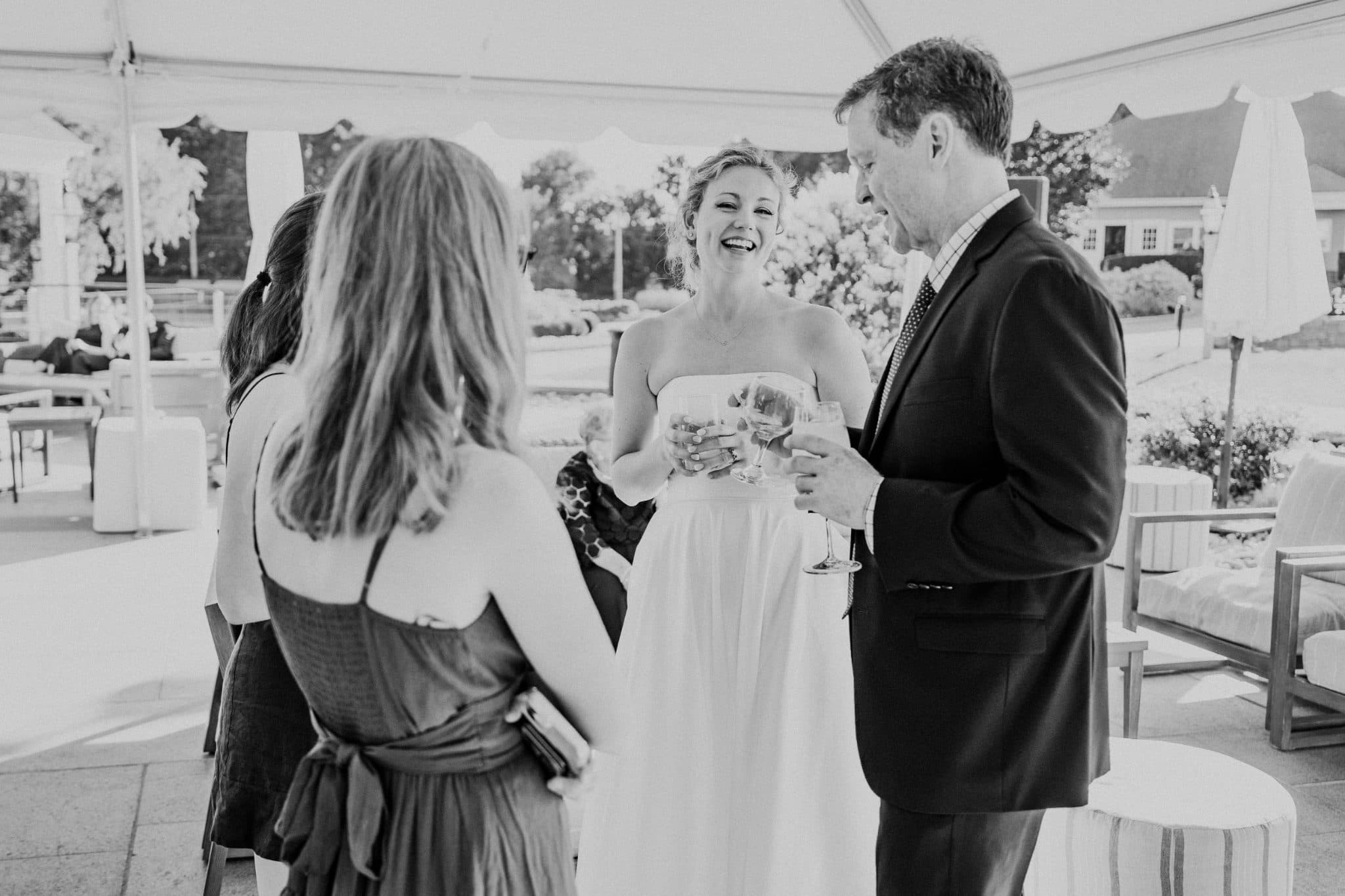 A bride in a strapless gown and a man in a suit hold glasses, smiling at each other in a tented outdoor setting.