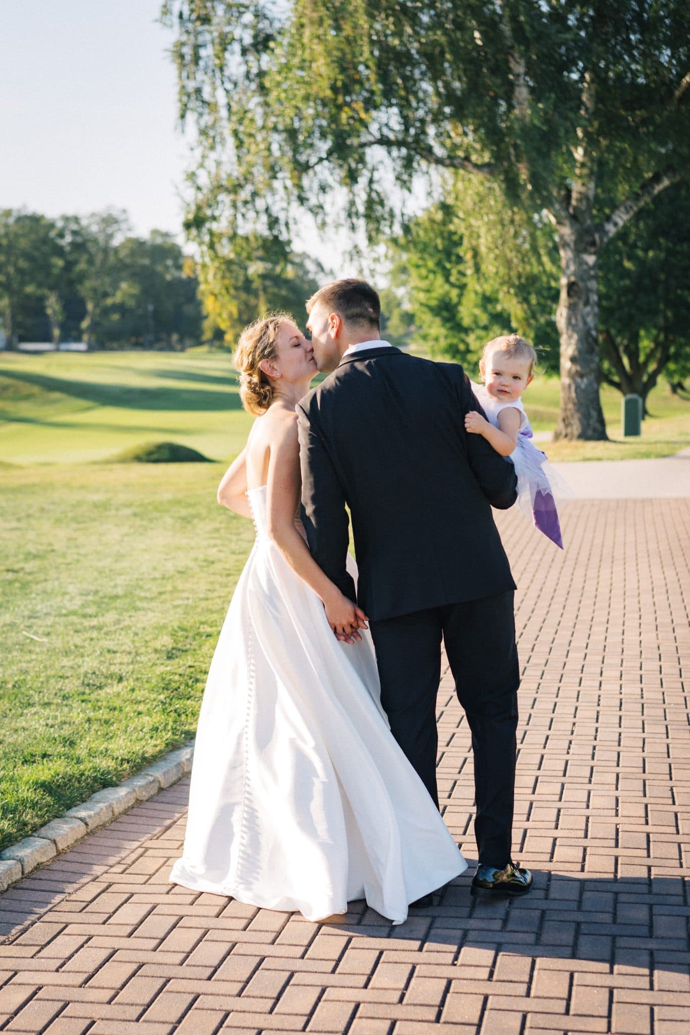 A bride in a white gown kisses a groom in a black suit while holding a baby in a white outfit on a brick path.