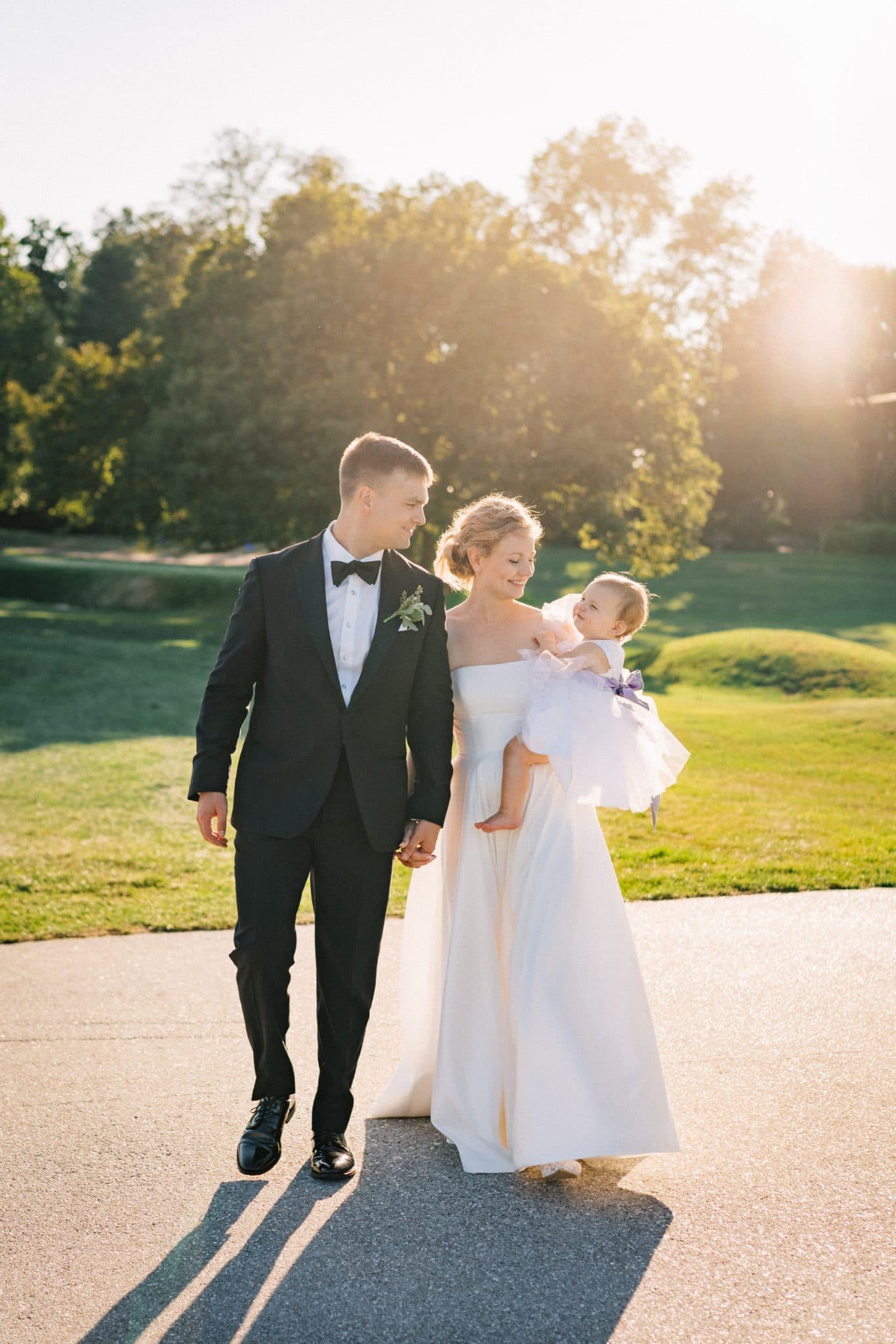 A bride in a white dress holds a baby, walking with a man in a black suit under a bright sky.