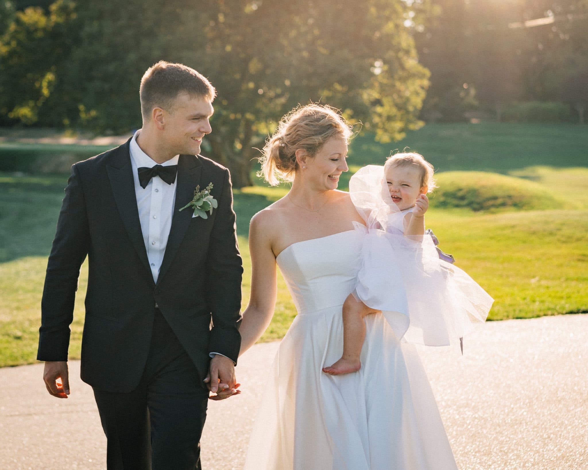 A bride in a white strapless gown holds a toddler in a white dress, while a groom in a black tuxedo and bow tie walks beside them.