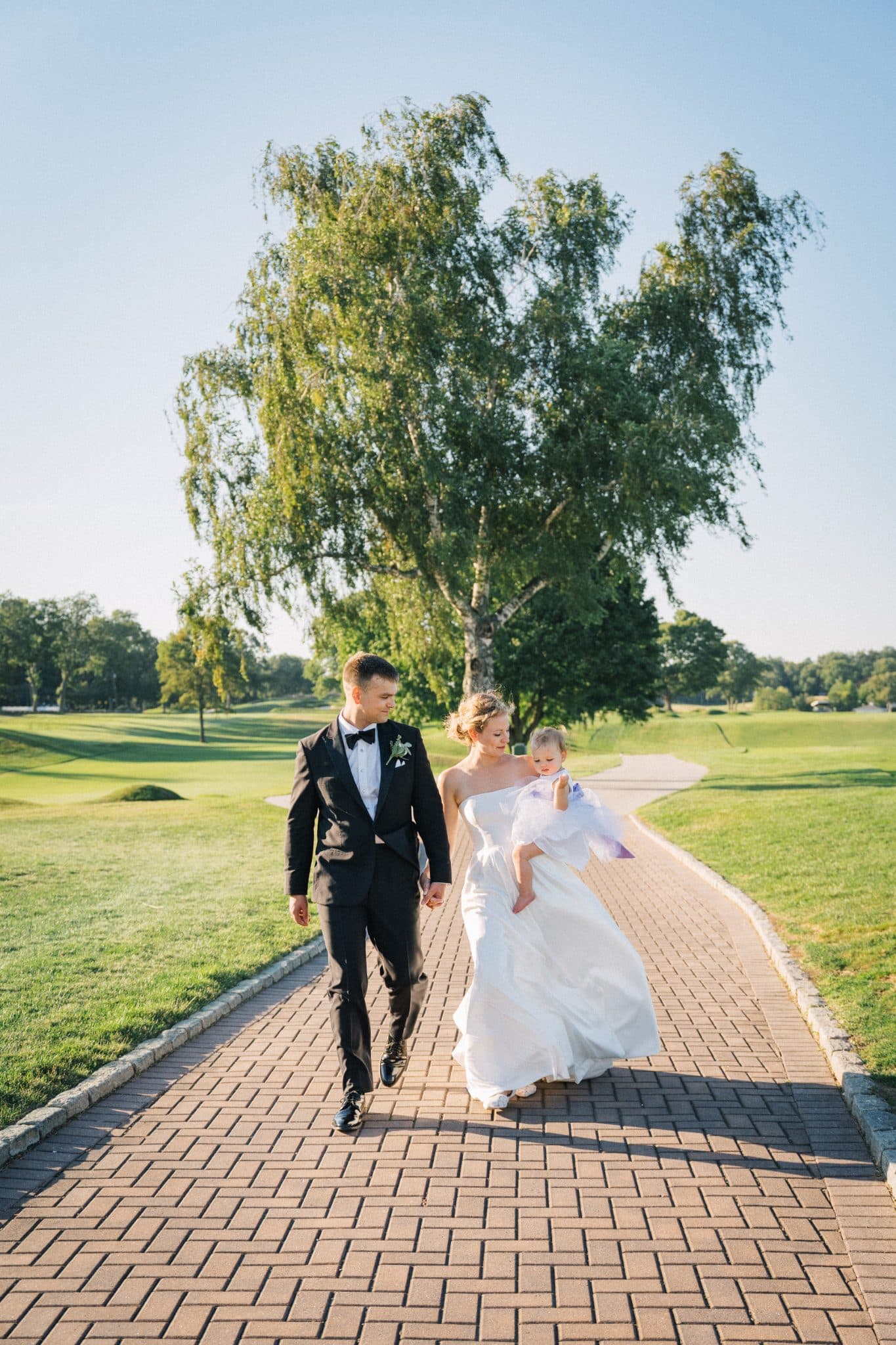 A bride in a white gown and a groom in a black suit walk hand in hand down a brick path, holding a small child.