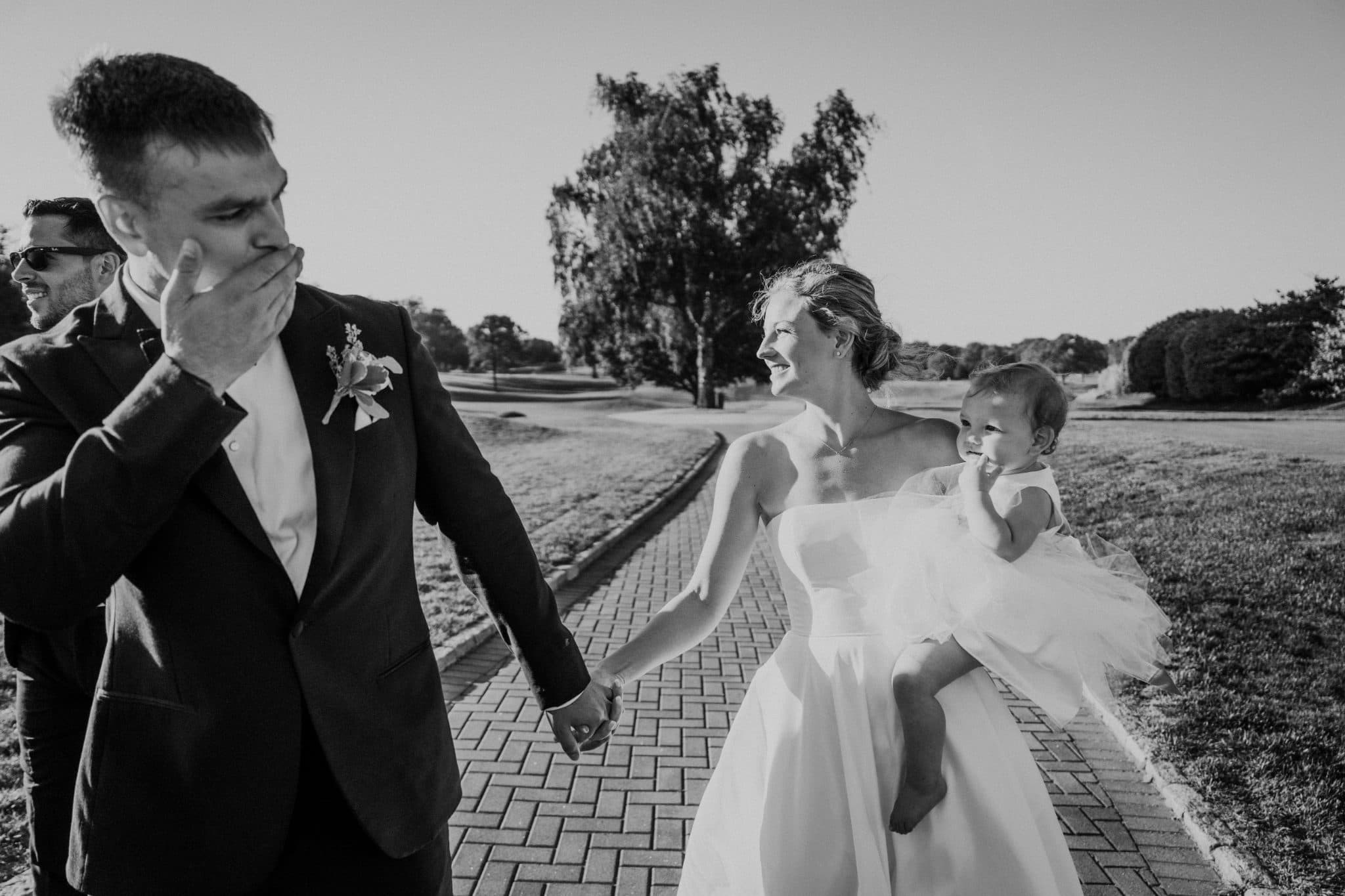 A bride in a white gown holds a baby while a man in a suit gestures with his hand.