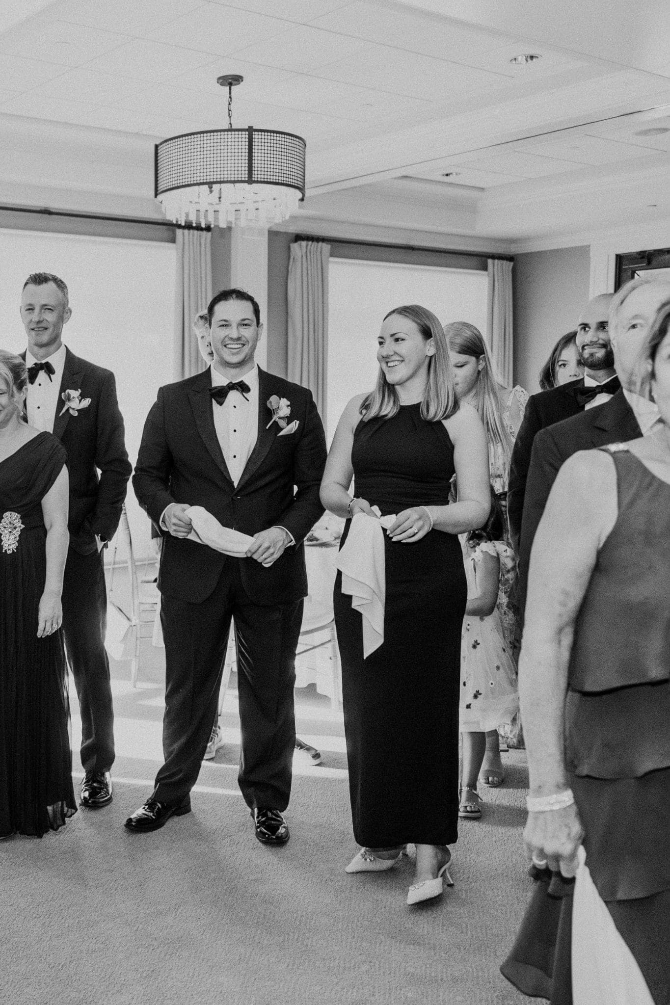 A black-and-white wedding scene shows a man in a tuxedo and a woman in a long dress walking together, surrounded by other guests in formal attire.