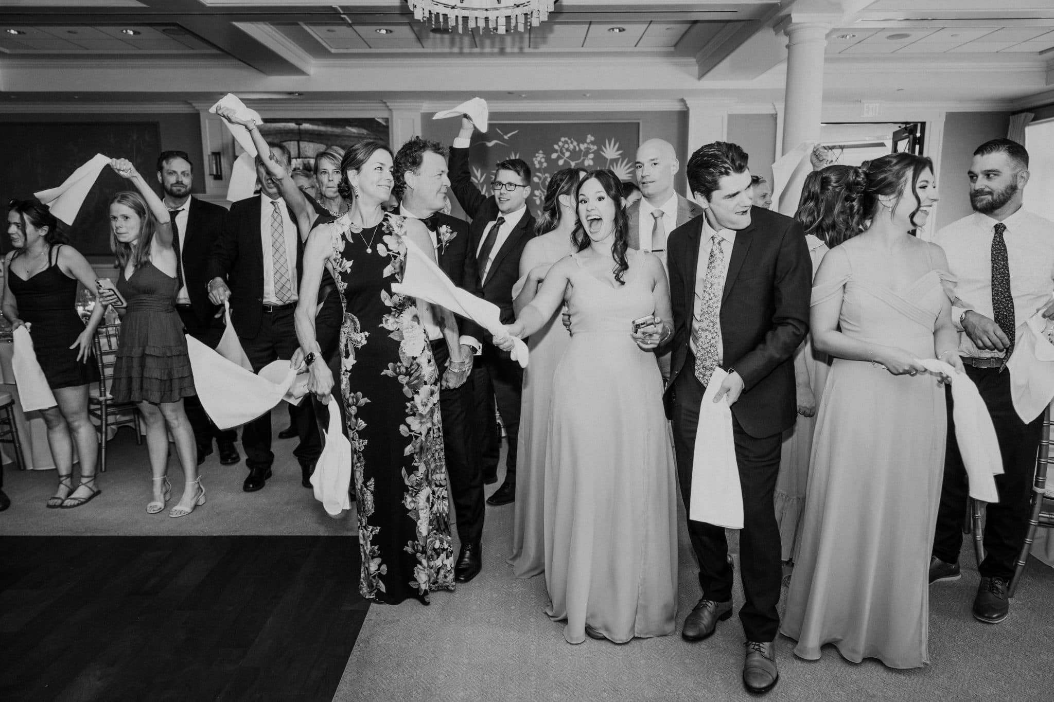 A group of people, including a bride in a light dress, are holding white napkins and celebrating in a room with a chandelier.