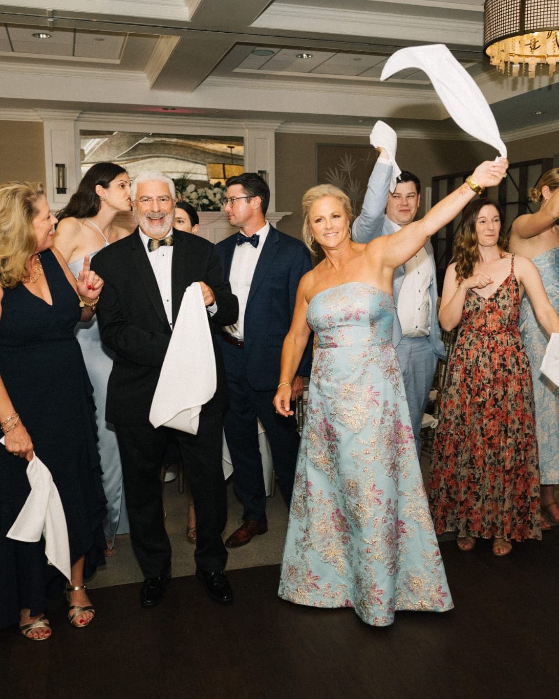 A group of people in formal attire holds white napkins, walking together in a room with chandeliers.