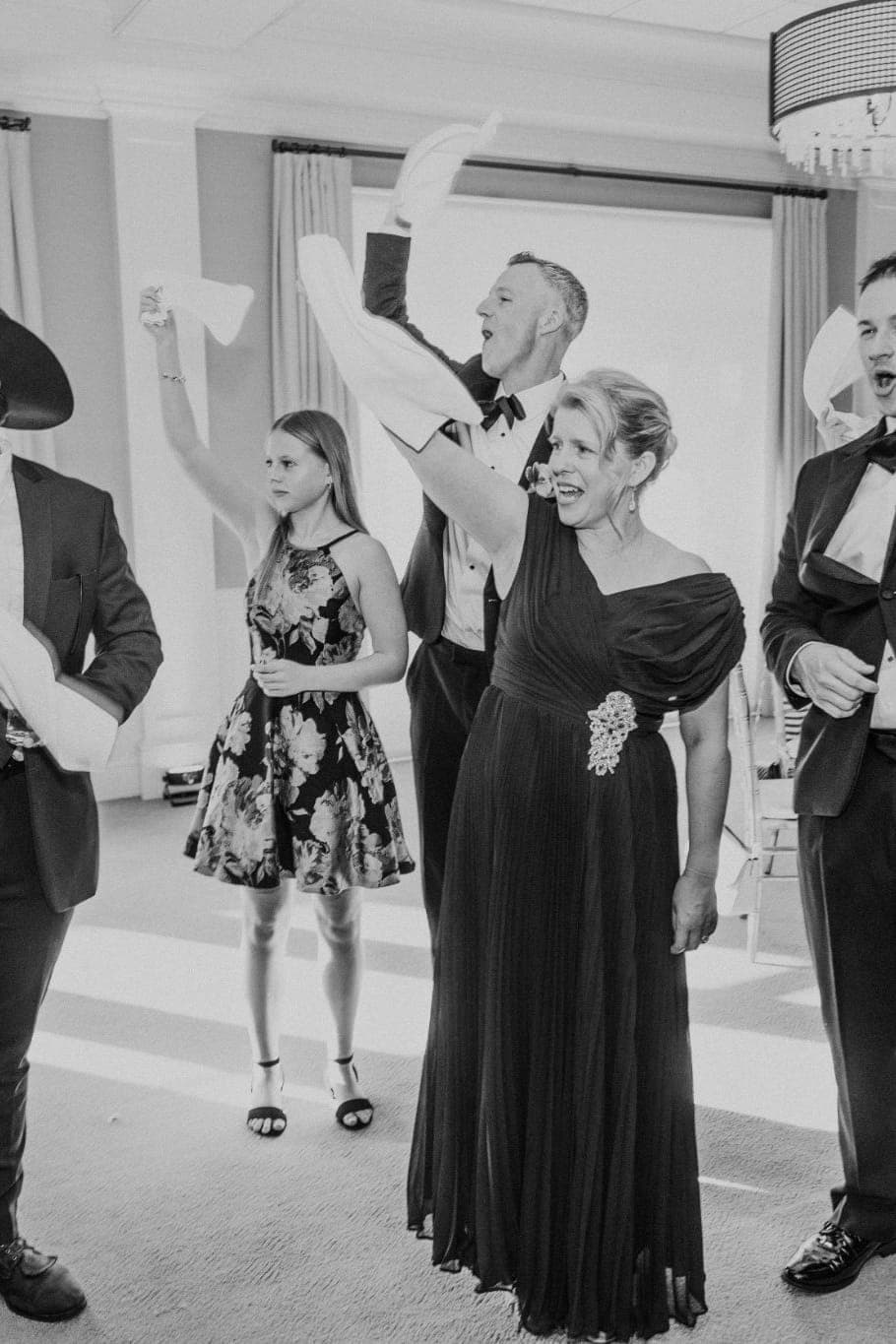 A group of people in formal attire stands in a room, some raising their arms, while one person holds a piece of paper.