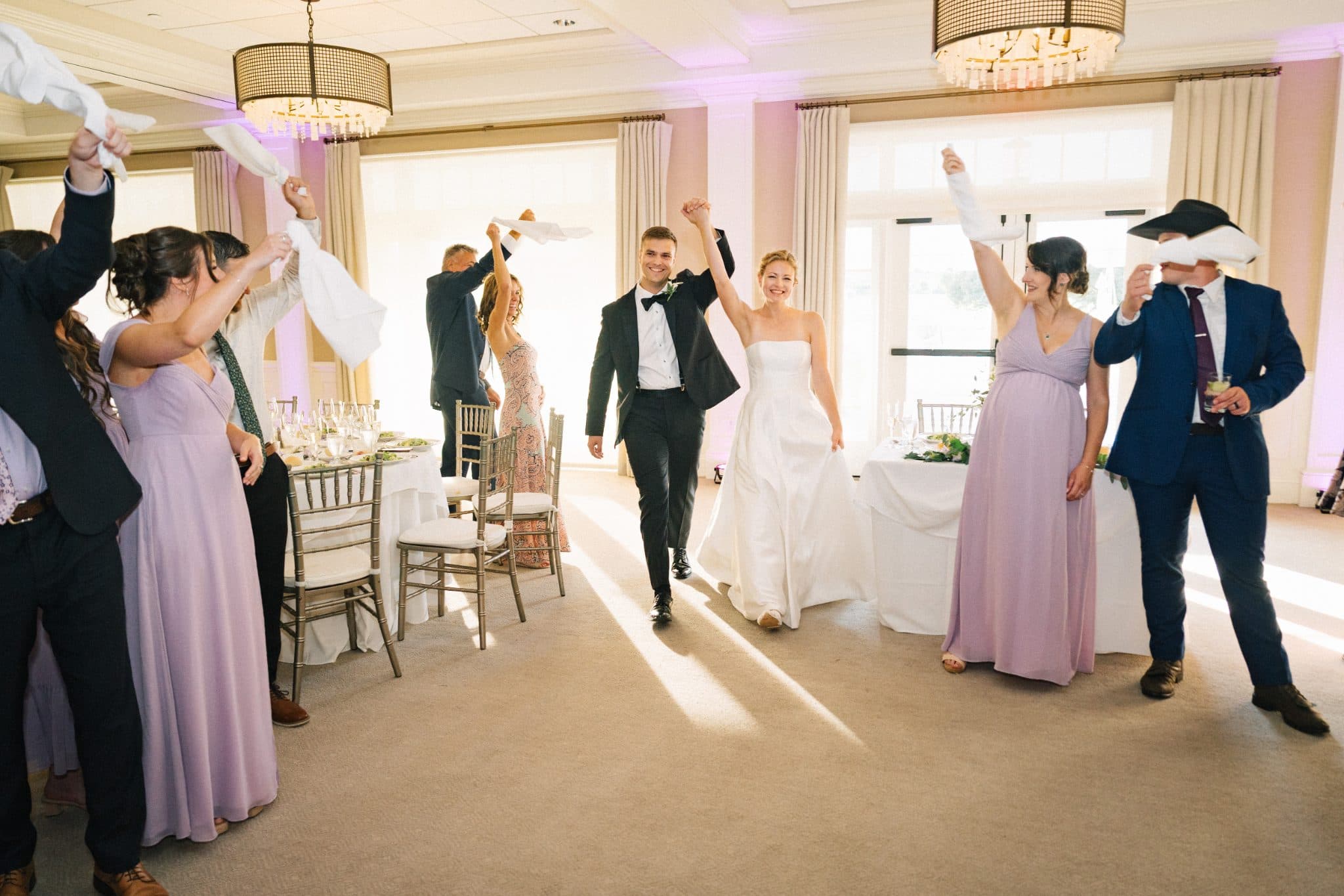 A bride and groom walk hand-in-hand through a room filled with guests raising white napkins in celebration.