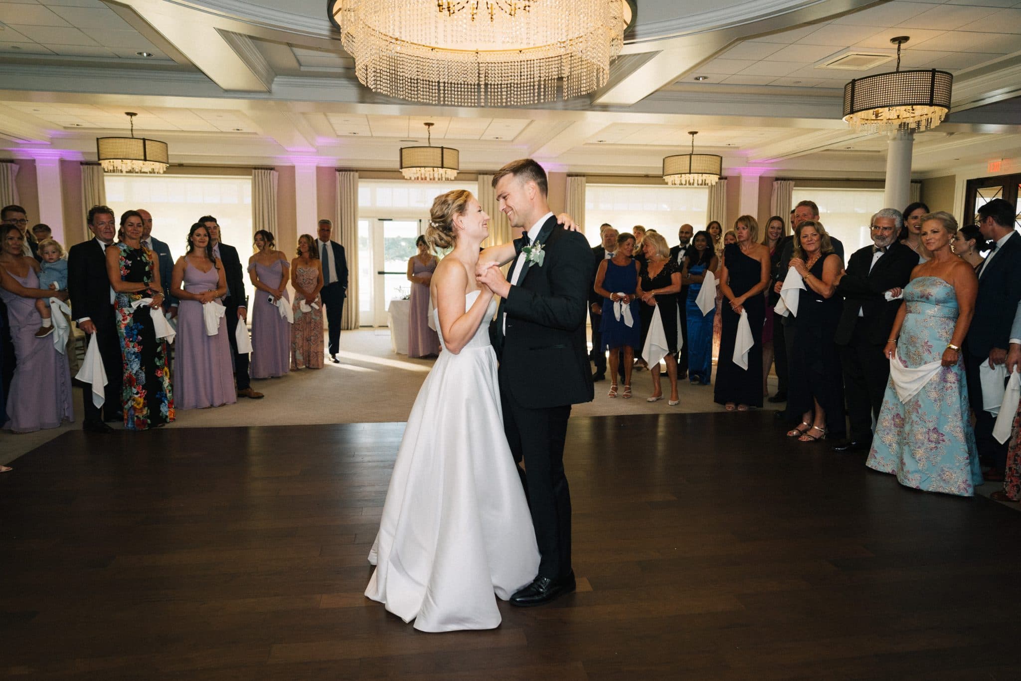 A bride and groom dance together on a wooden floor, surrounded by guests in formal attire.