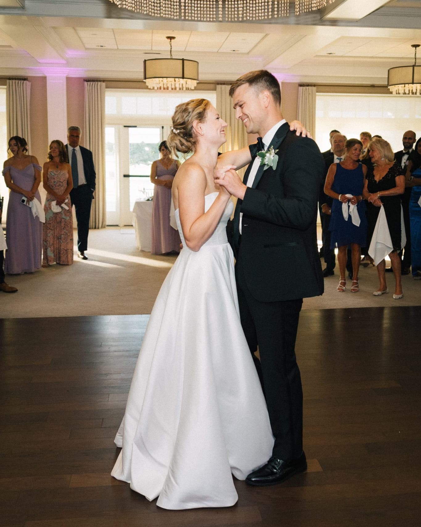 A bride in a white gown dances with a man in a black suit under a chandelier in a well-lit room.