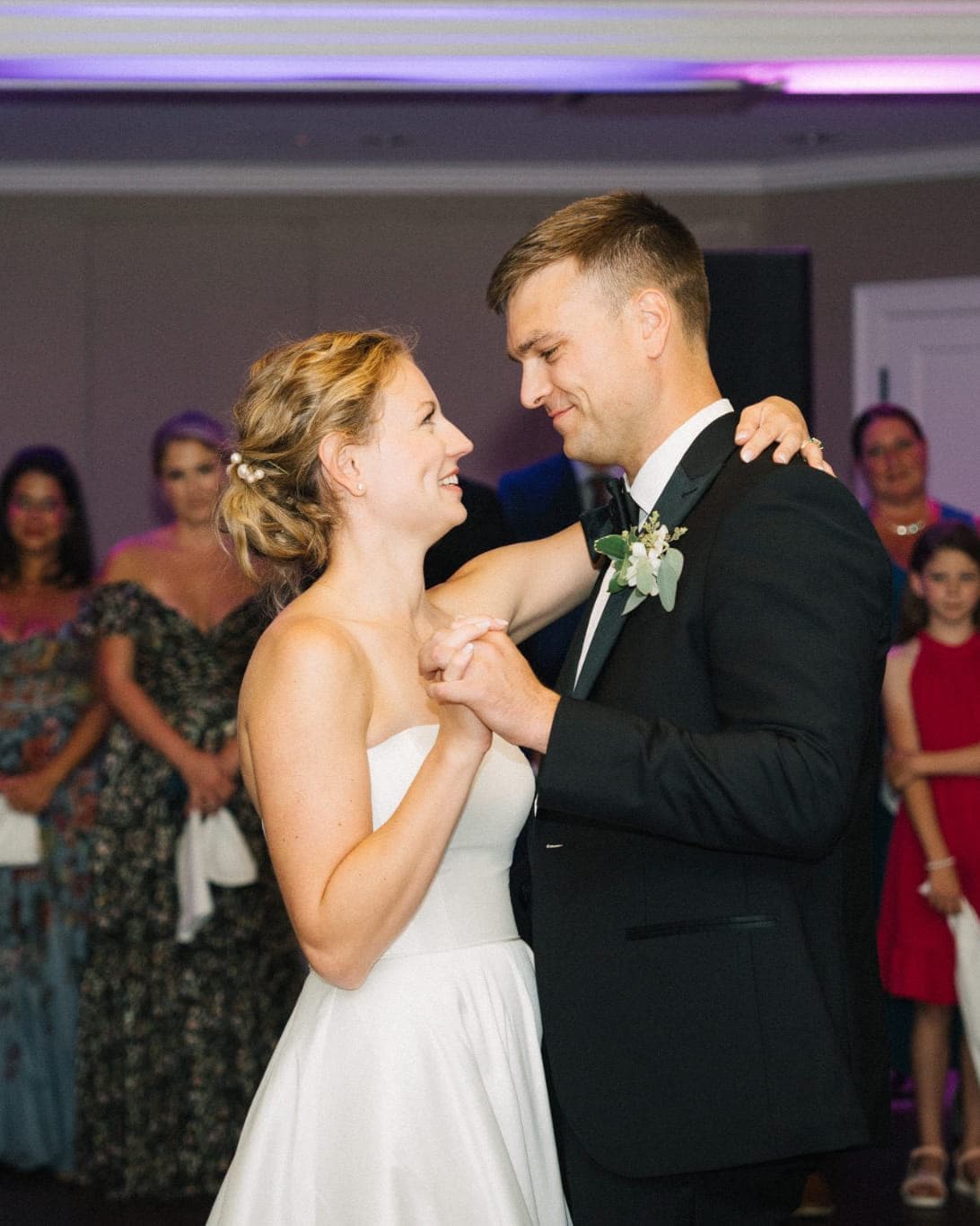 A bride and groom dance together at a wedding reception, surrounded by guests in formal attire.
