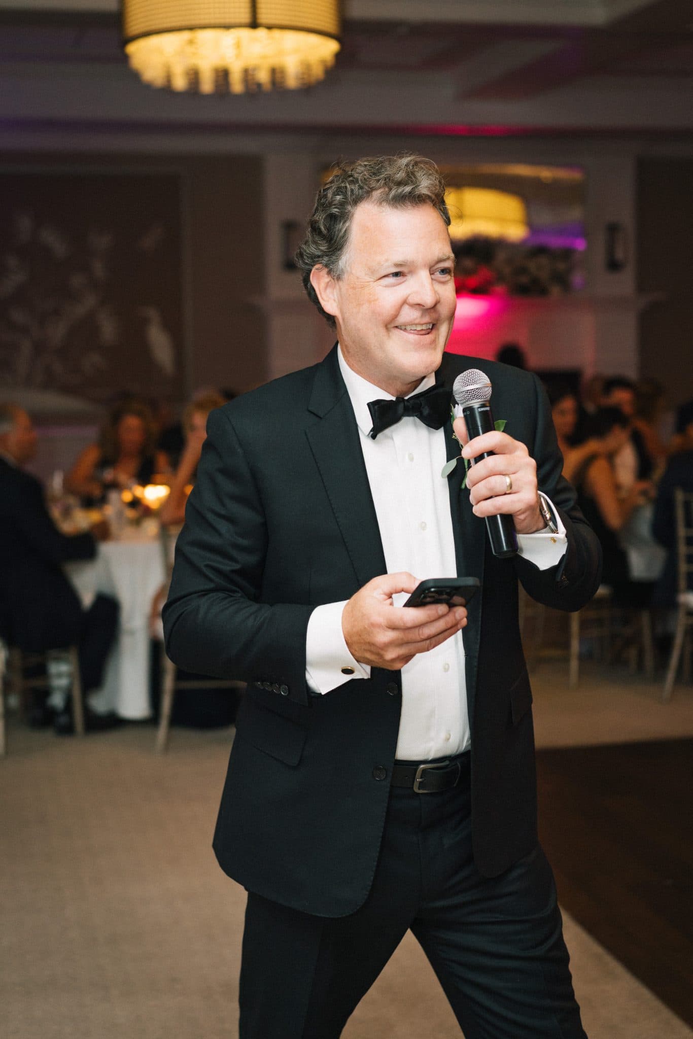 A man in a tuxedo holds a microphone and a phone, smiling in a warmly lit room with guests seated at tables.