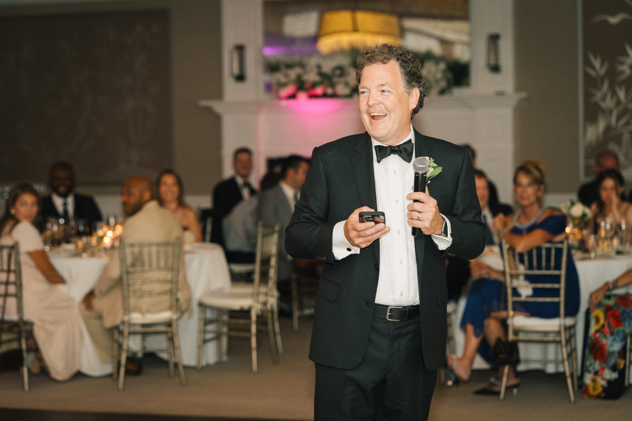 A man in a tuxedo holds a microphone and a phone, smiling at a wedding reception with guests seated at tables.