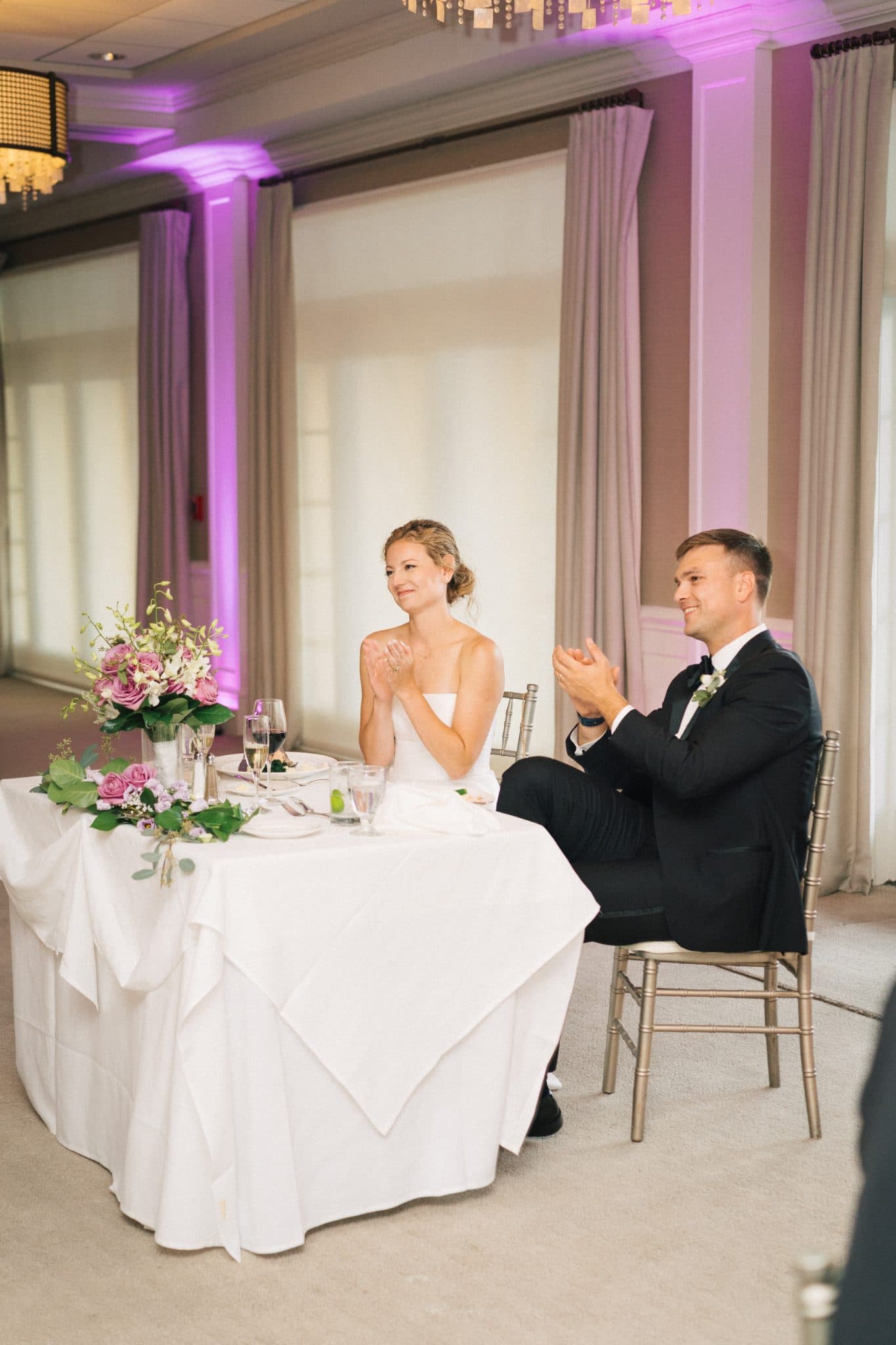 A bride and groom sit at a table with a white tablecloth, clapping, surrounded by floral decorations and elegant decor.