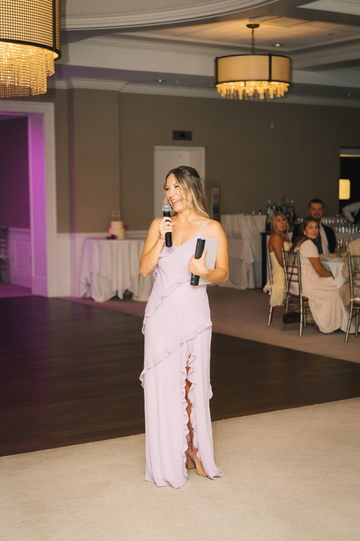 A woman in a lavender gown holds a microphone and a notepad, standing on a dance floor in a well-lit room with guests seated at tables.