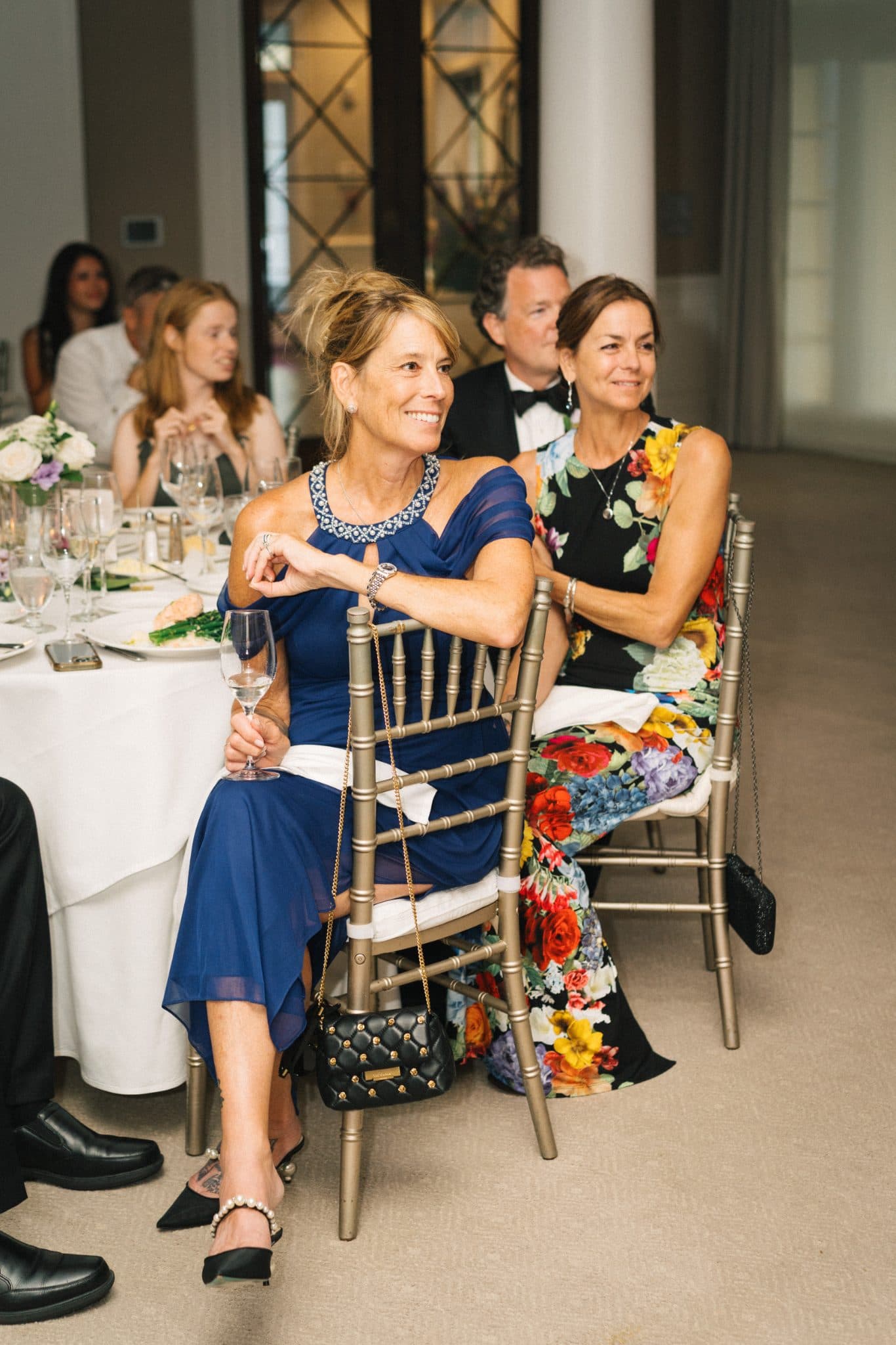 Two women in elegant dresses sit at a table with wine glasses, smiling and looking to the side.