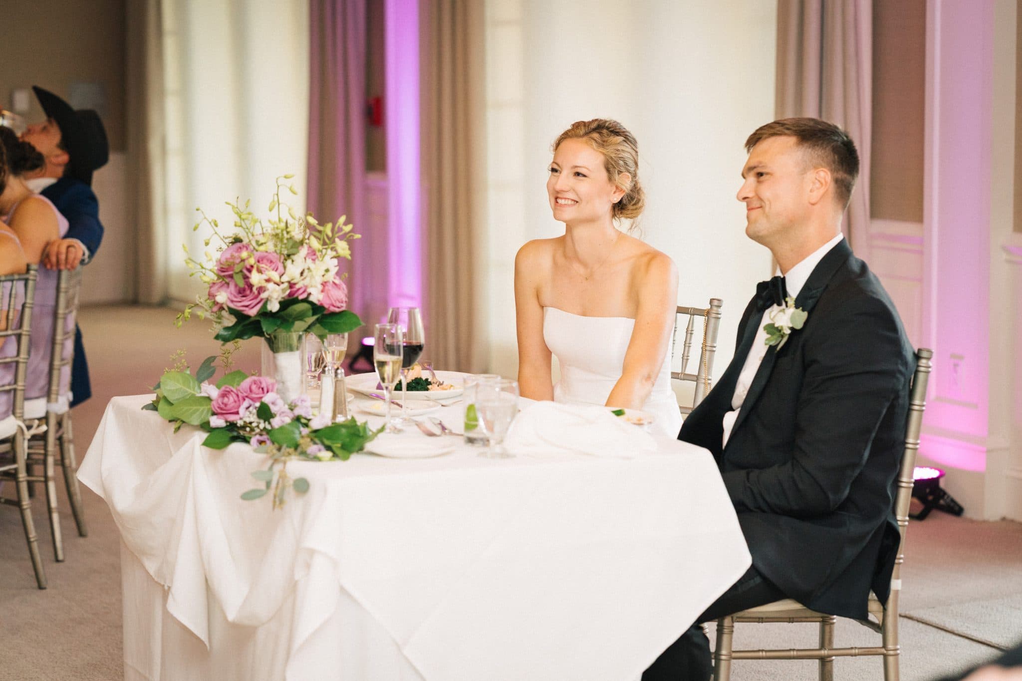 A bride in white and groom in black suit sit at a table with pink flowers and white tablecloth.