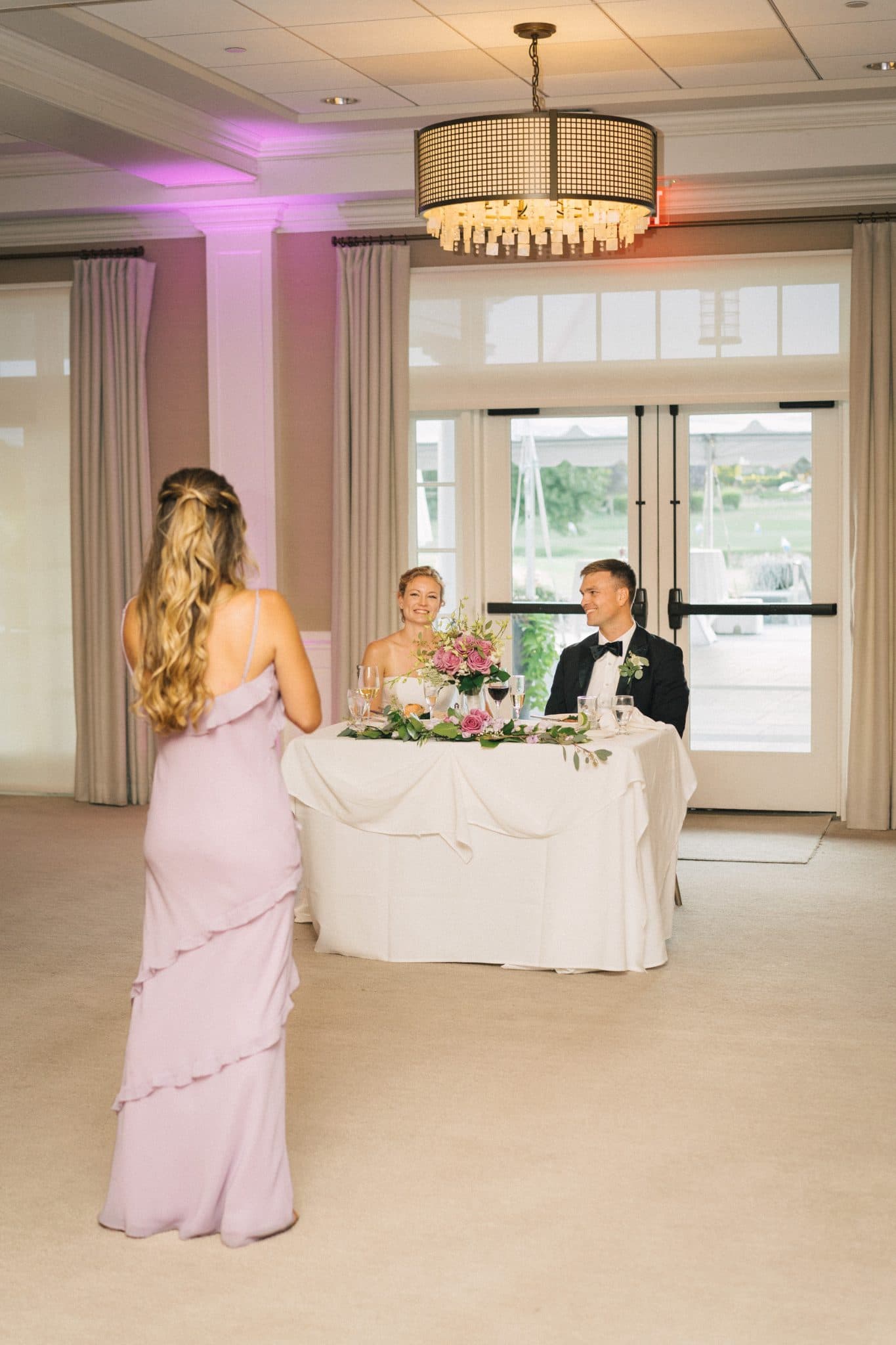A woman in a lavender gown stands facing a couple seated at a table adorned with flowers and wine glasses.