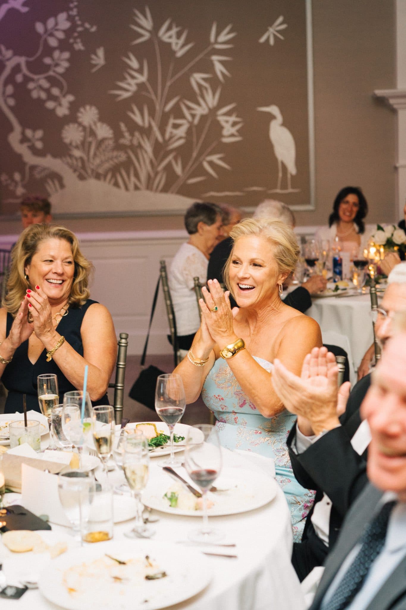 At a wedding reception, guests at a table clap and smile, surrounded by plates of food and wine glasses.
