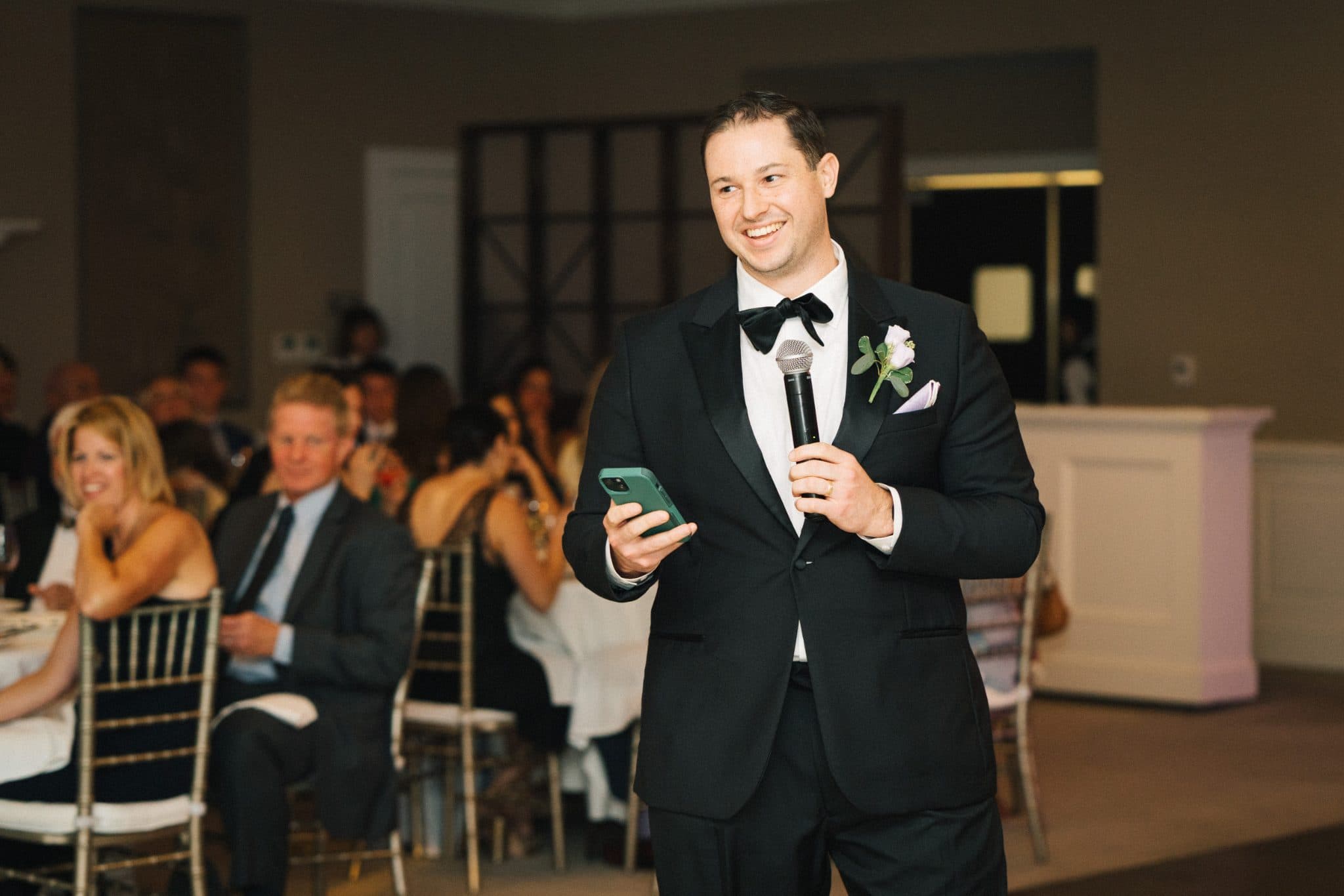 A man in a tuxedo holds a microphone and a phone, smiling at a seated audience in a banquet hall.