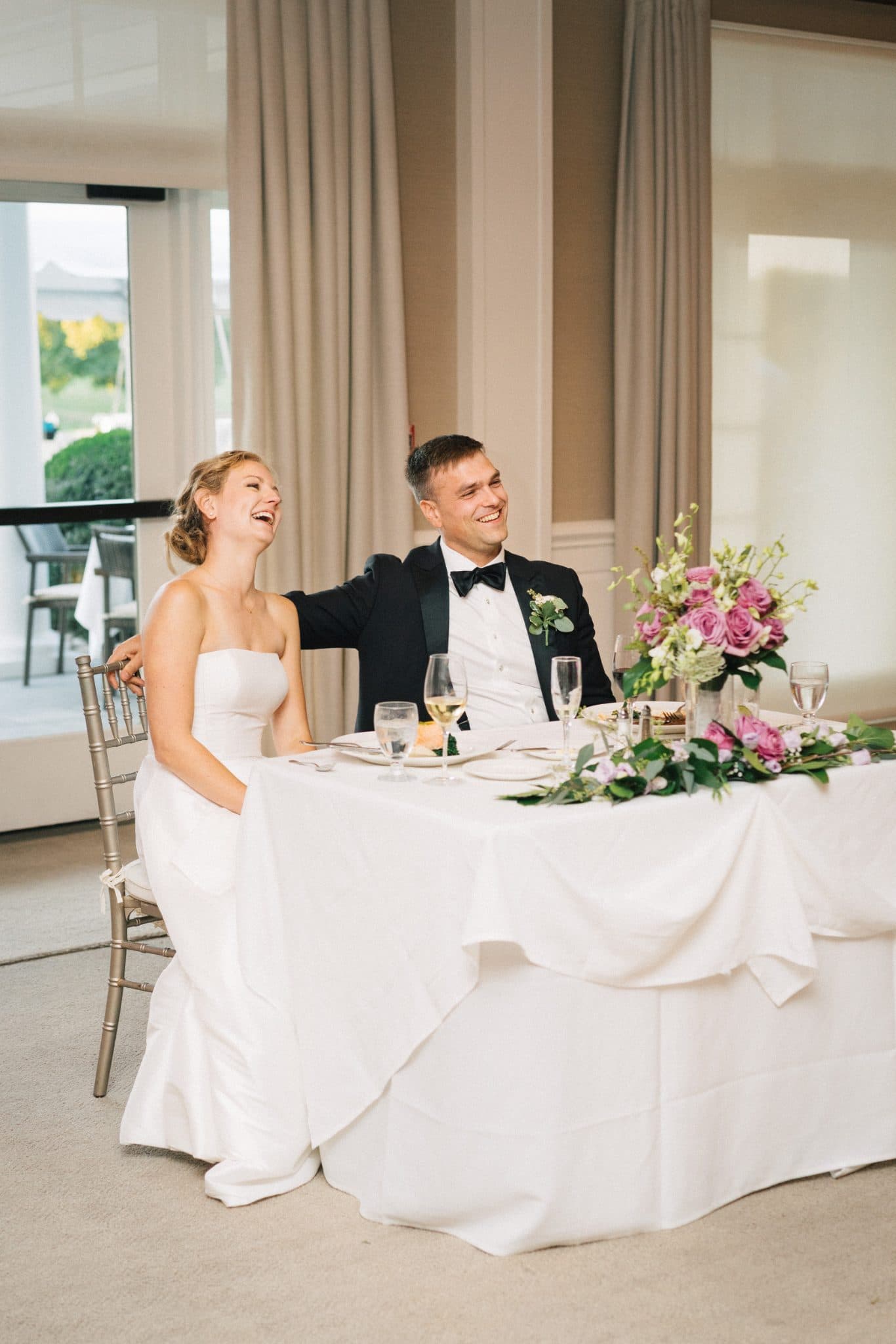 A bride in a white gown and a groom in a tuxedo sit at a white table with a floral centerpiece, laughing and enjoying the moment.