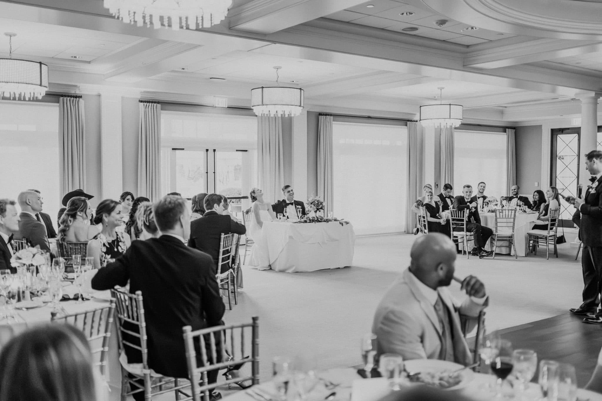 A black and white wedding reception with guests seated at tables, a bride and groom at the head table, and a man standing at the podium.