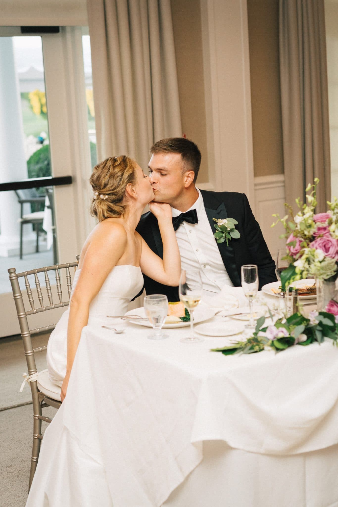 A bride and groom share a kiss at their wedding reception table adorned with flowers and wine glasses.