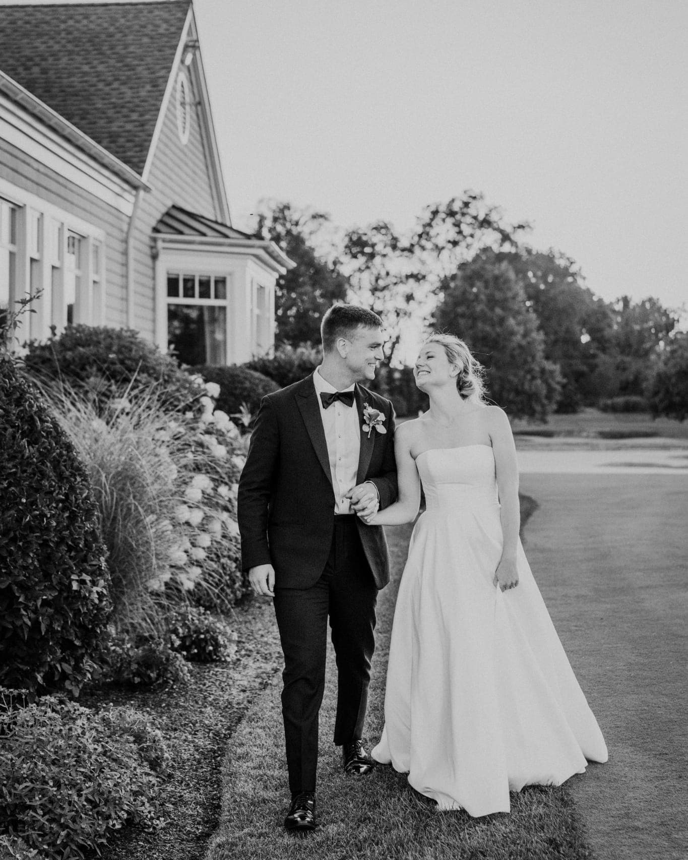 A bride in a strapless gown and a groom in a tuxedo walk hand in hand on a grassy path near a building with a sloped roof.