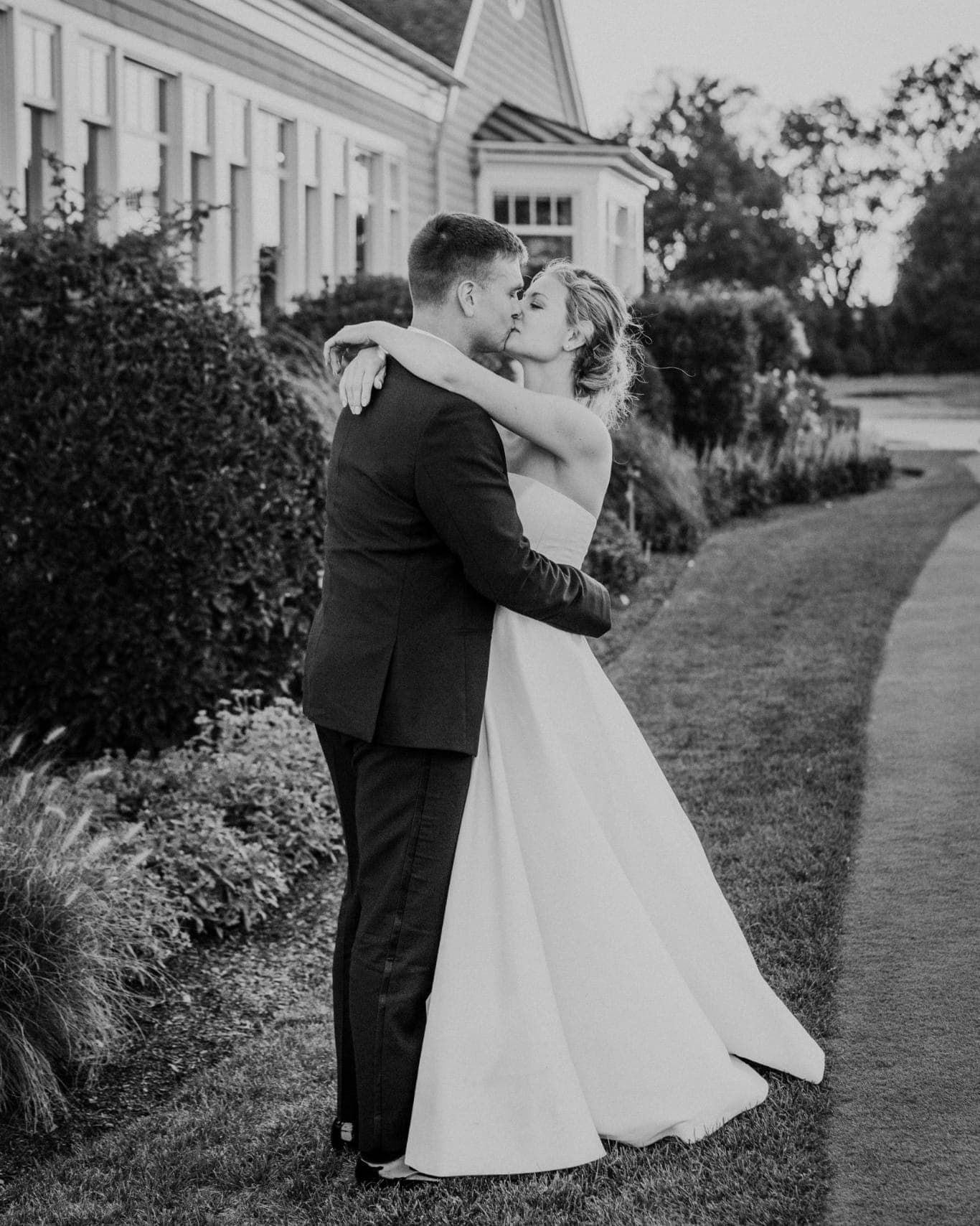 A bride in a white gown and groom in a dark suit share a kiss on a grassy path near a building with a sloped roof.