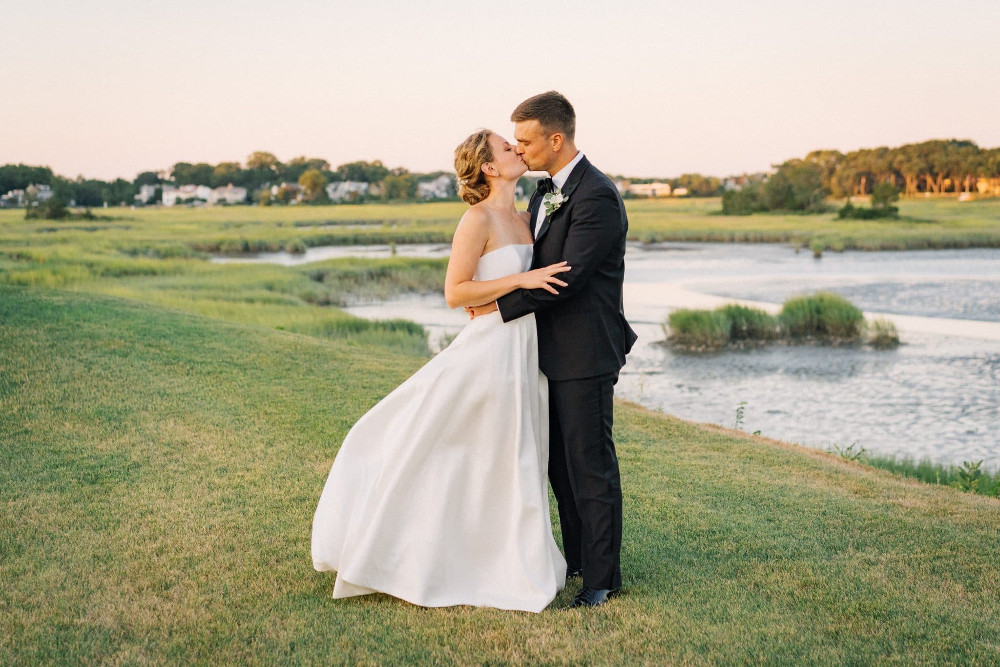 A bride and groom share a kiss on a grassy hillside overlooking a marshy area.