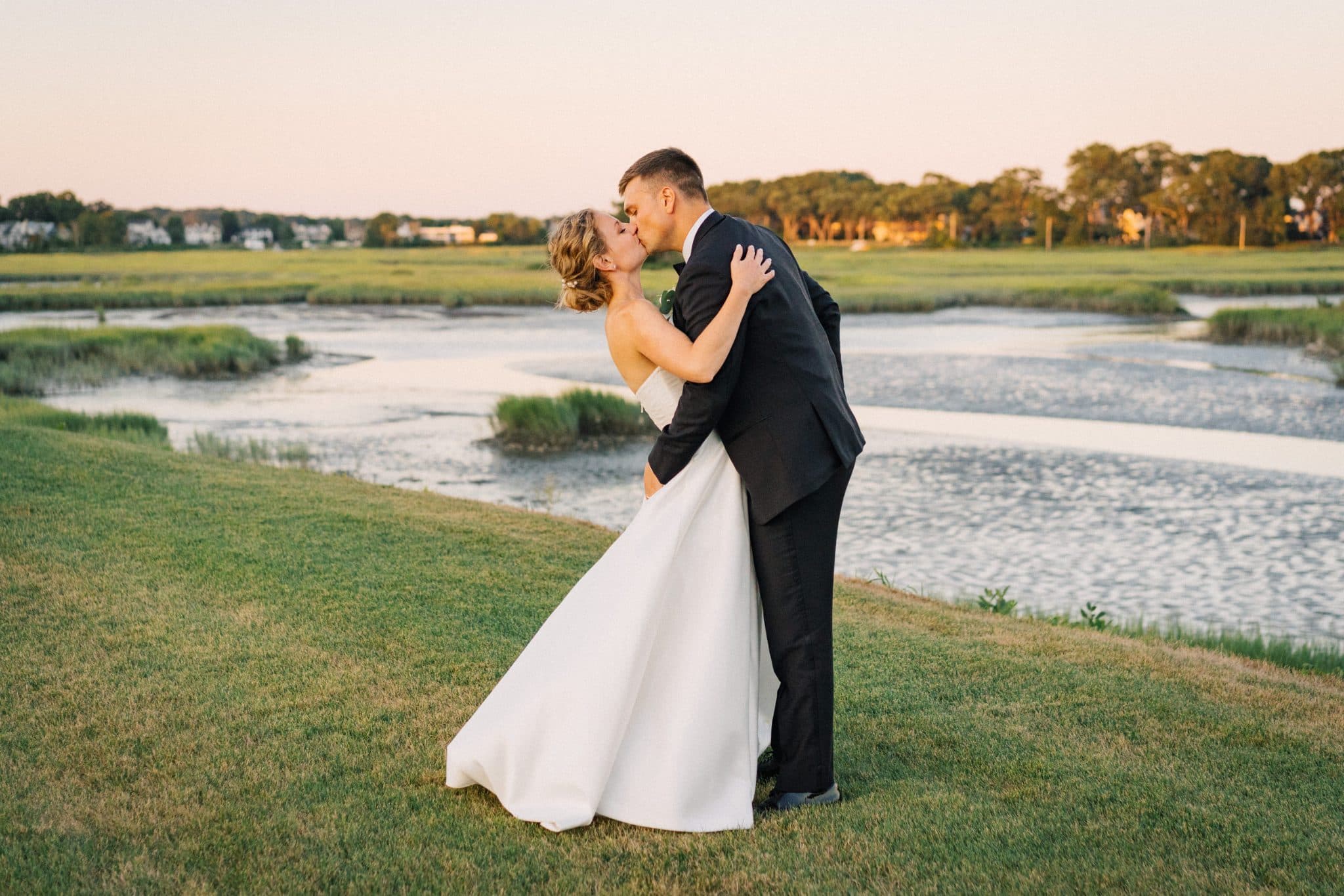 A bride and groom share a kiss on a grassy hill overlooking a river and trees.