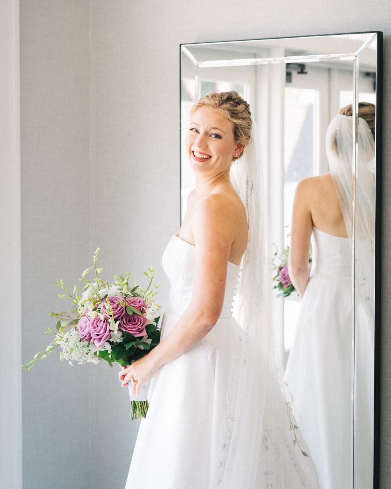 A bride in a white dress holds a bouquet of purple and white flowers, smiling at the mirror.