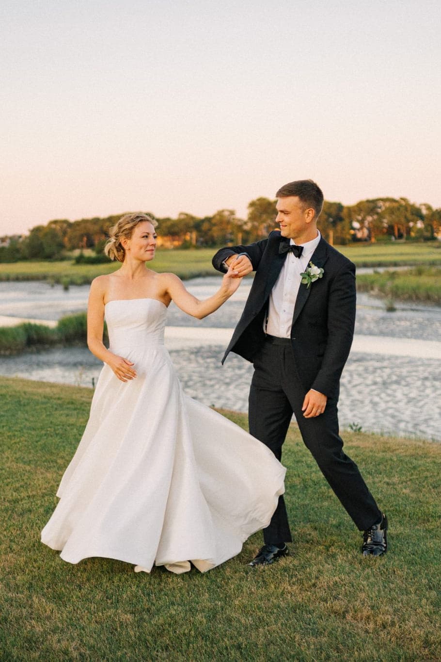 A bride in a strapless white gown and a groom in a black tuxedo hold hands, walking on a grassy hillside with a river and trees in the background.