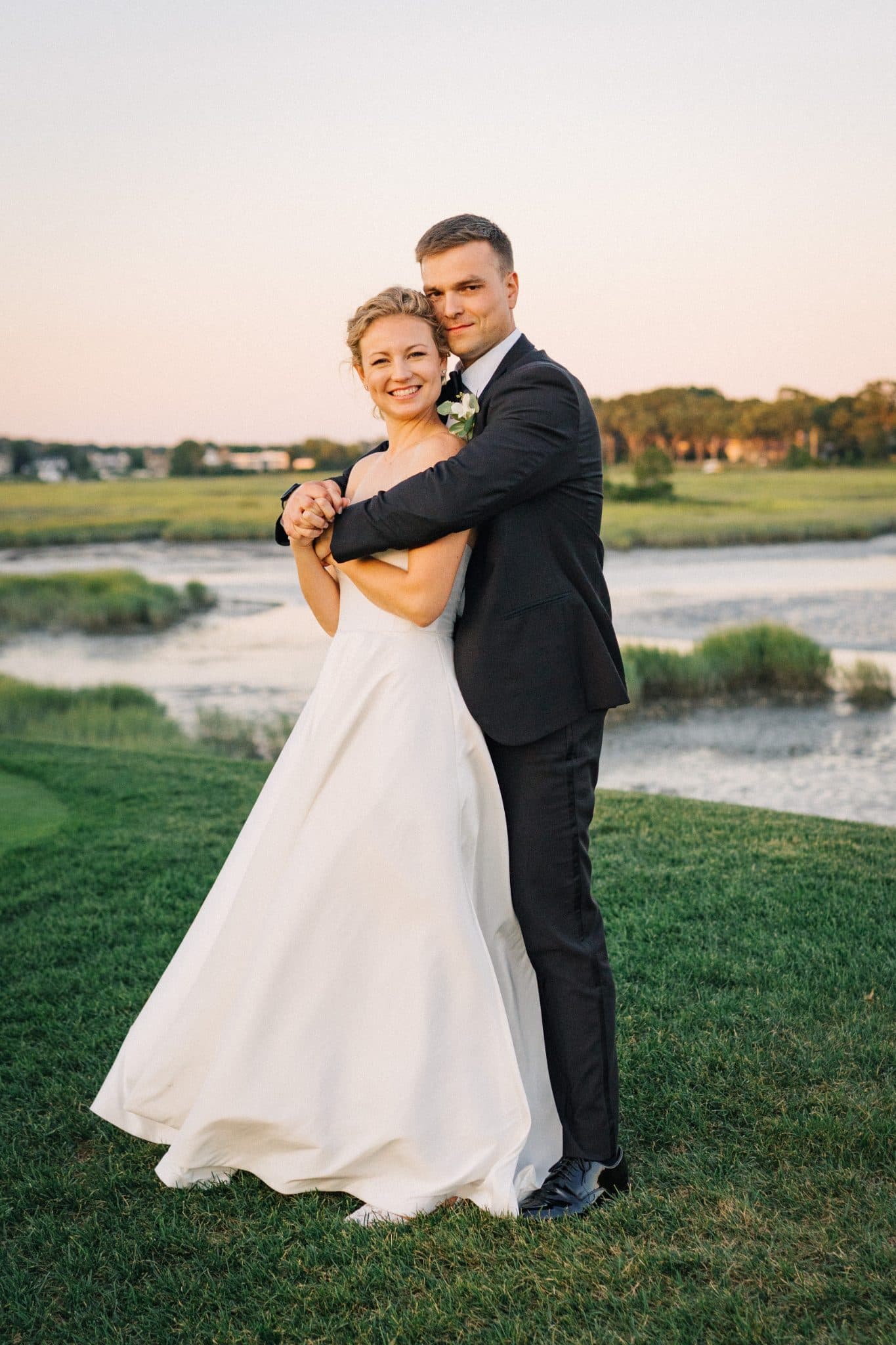 A bride in a white gown and a groom in a dark suit embrace on a grassy field near a body of water.