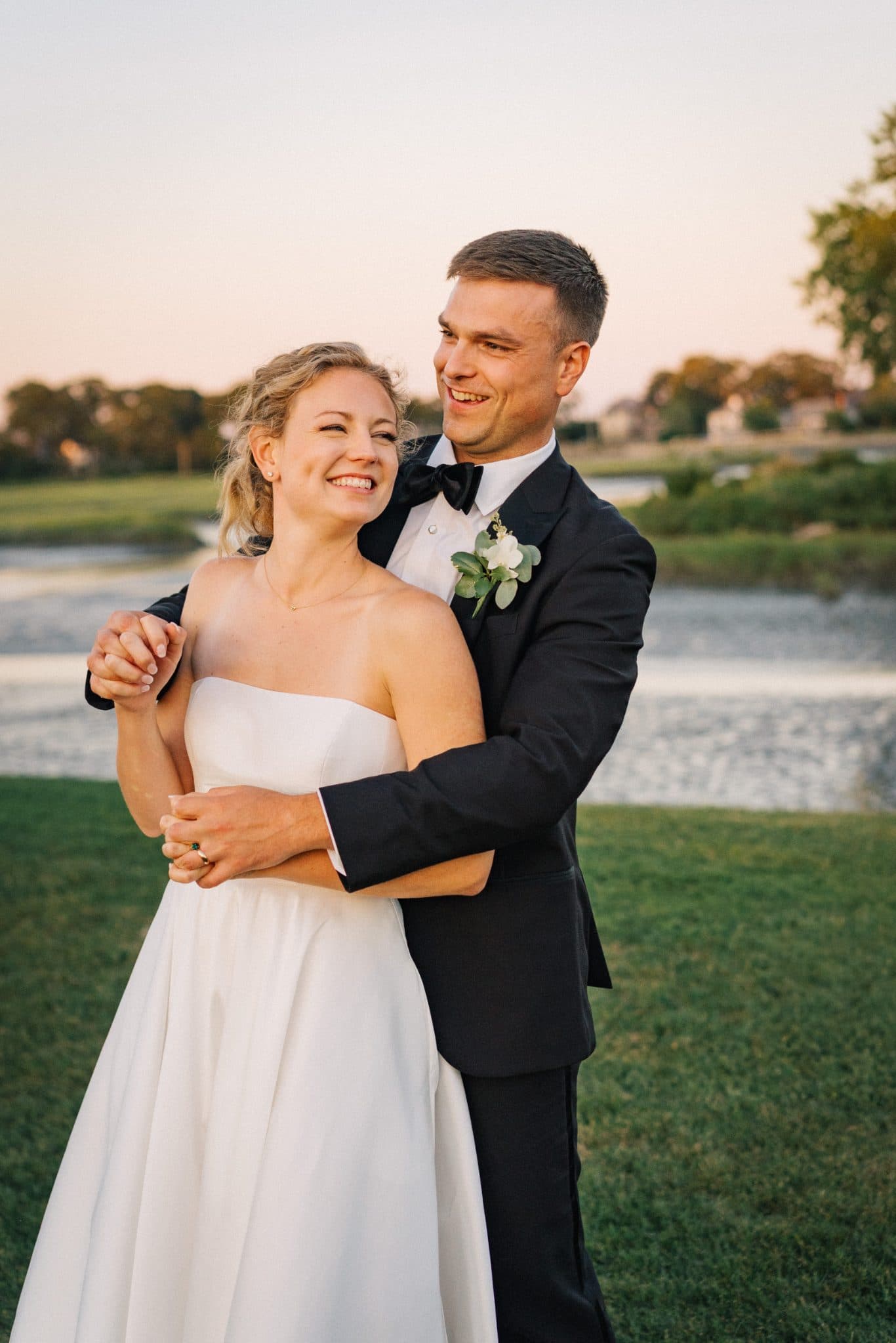A bride in a strapless white gown and a groom in a black tuxedo embrace in a grassy area near a body of water.