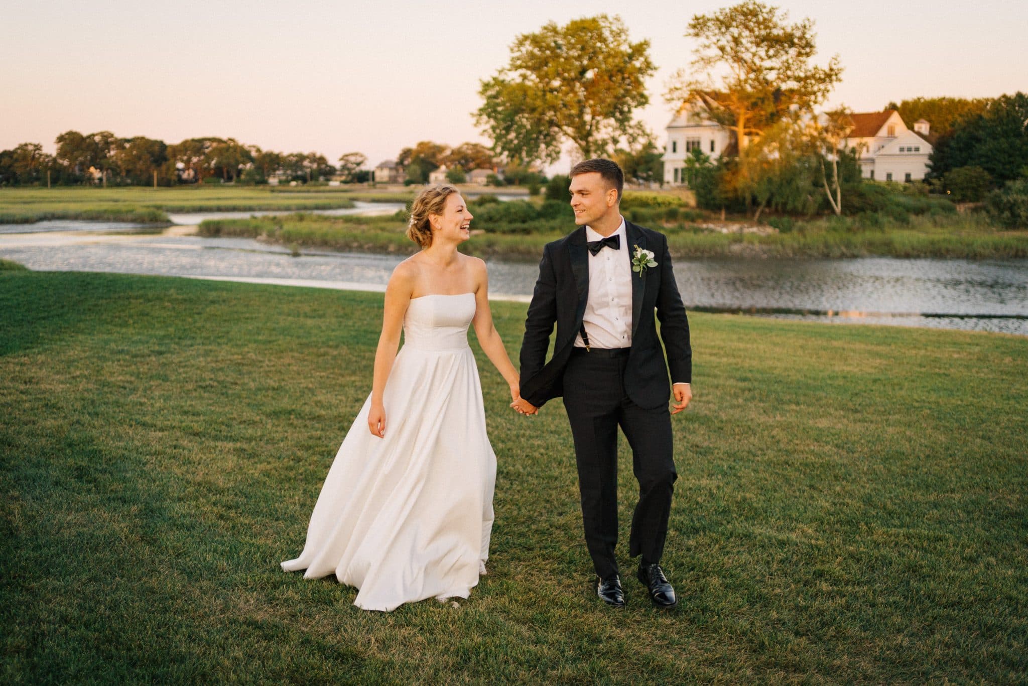 A bride in a white strapless gown and a groom in a black tuxedo walk hand in hand on a grassy field near a body of water and trees.
