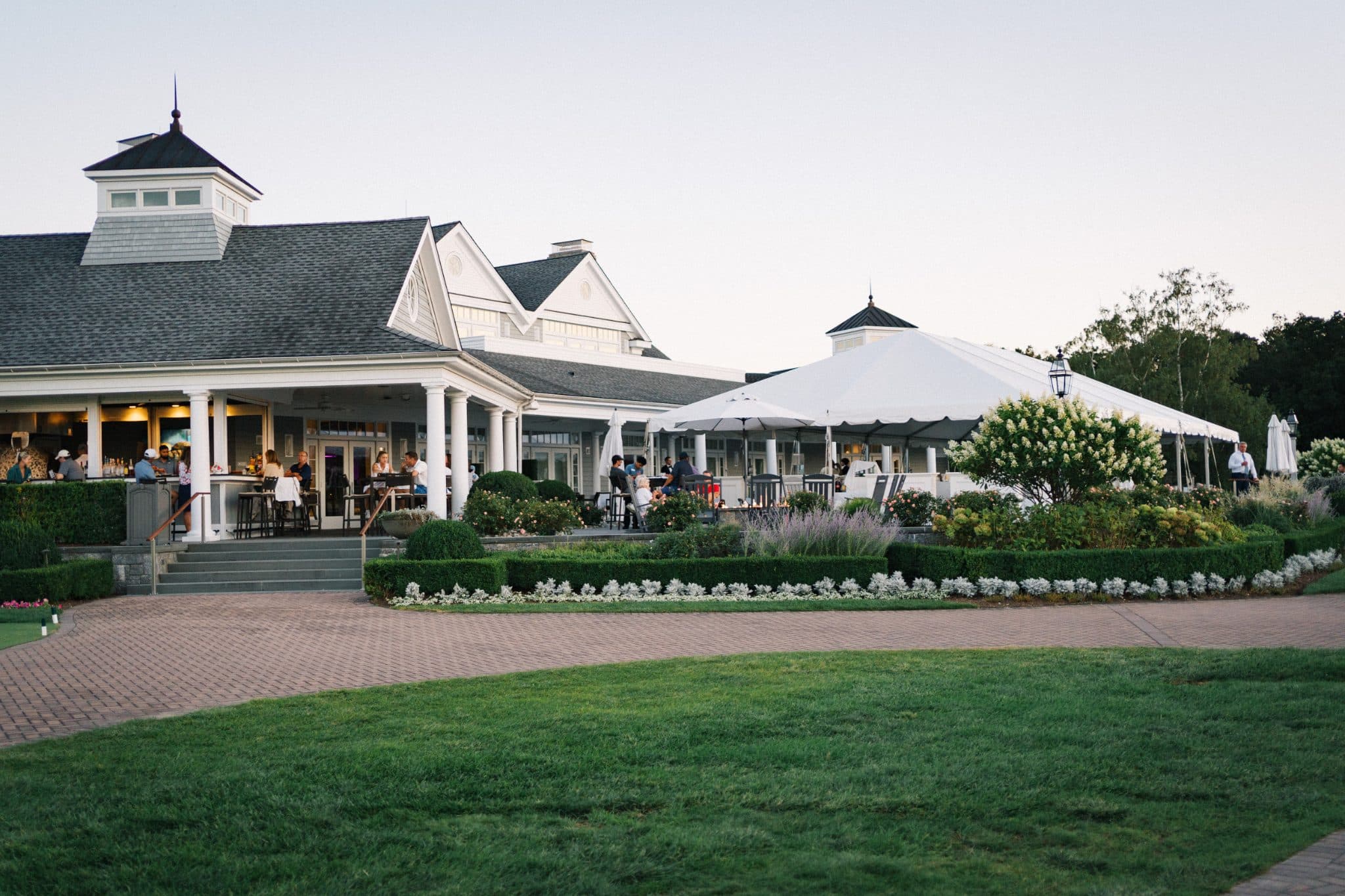 A white tent with people gathered under it, surrounded by a well-maintained garden and a building with a porch.