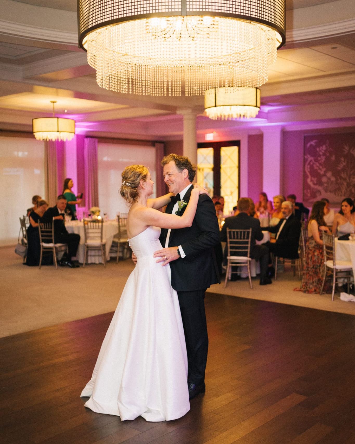 A bride in a white gown dances with a man in a black suit under a large chandelier in a warmly lit room.