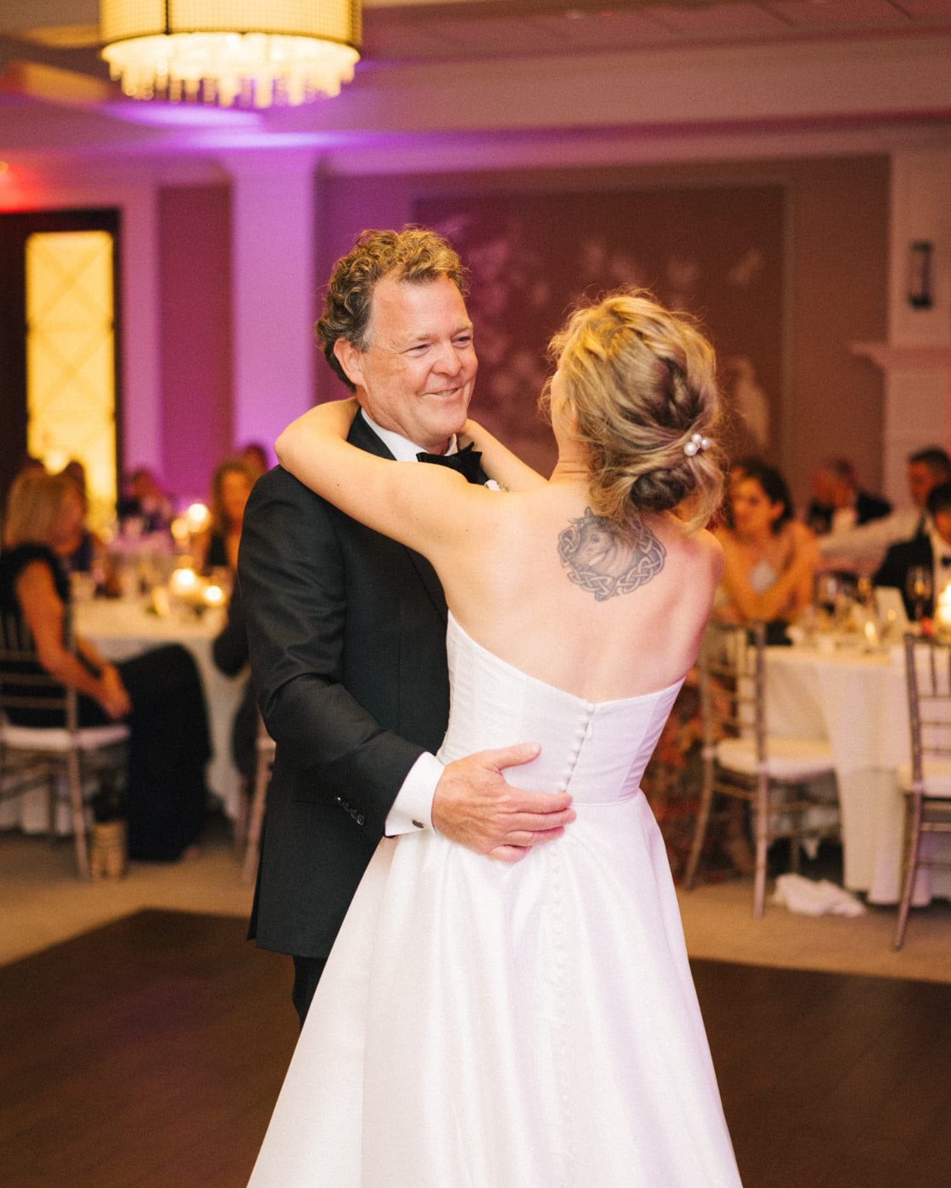 A bride in a white dress dances with a man in a tuxedo under a chandelier in a warmly lit room.