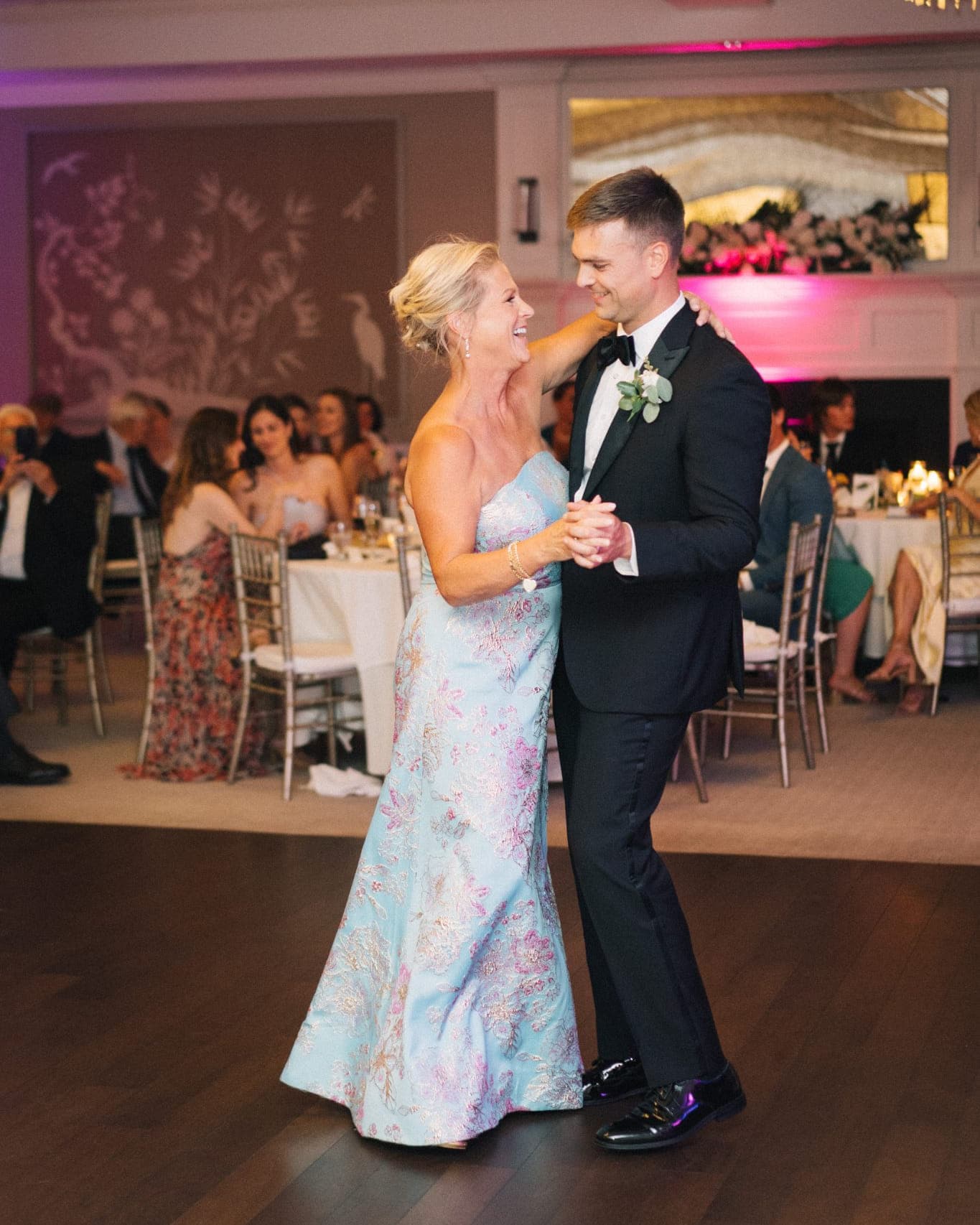 A couple dances on a wooden floor in a well-lit room with guests seated at tables in the background.