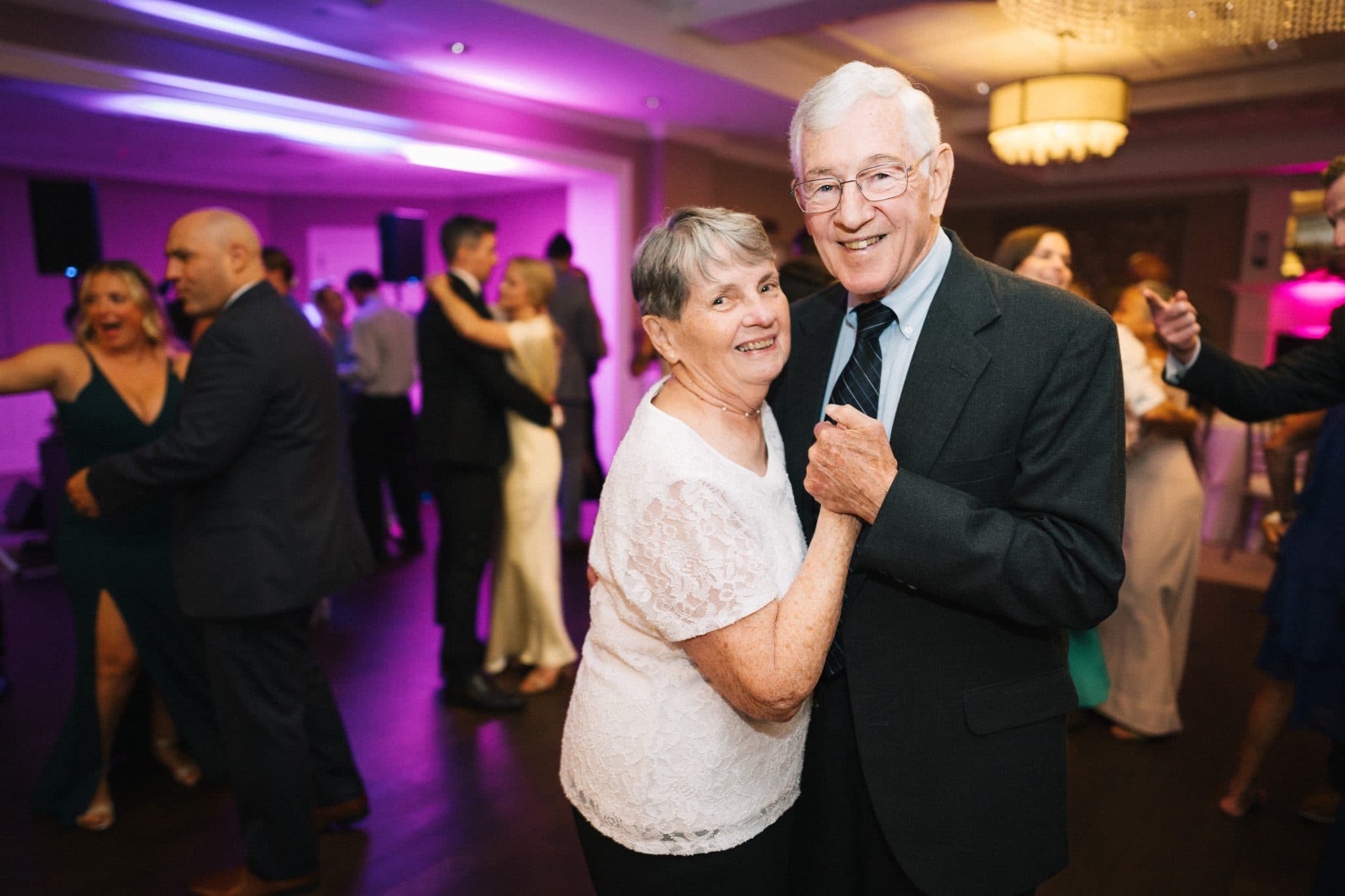 An older couple dances joyfully in a lively wedding reception with purple lighting.
