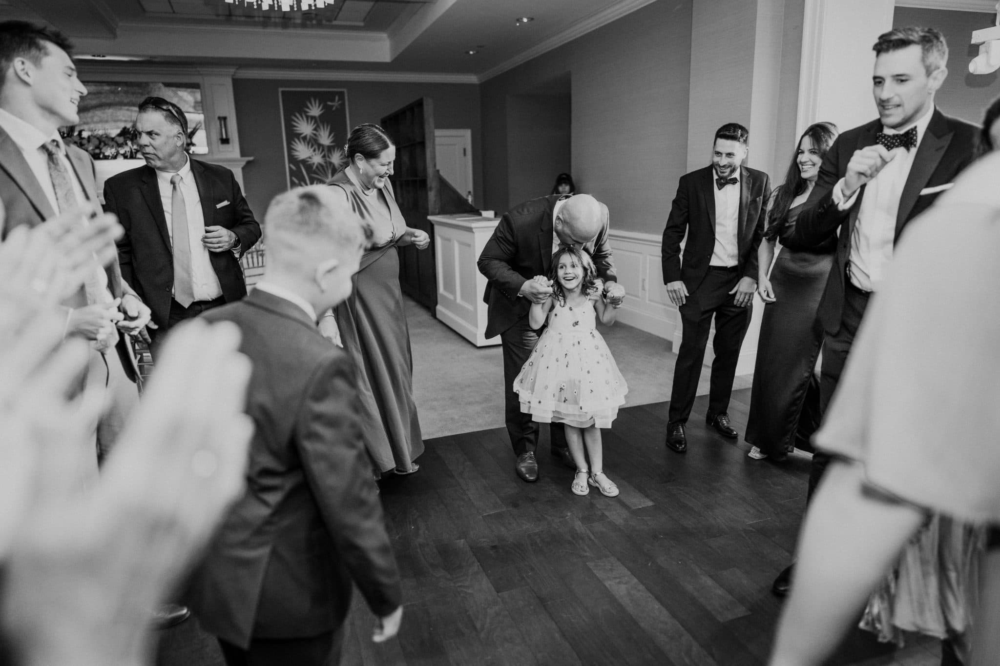 A group of people, including a child in a dress, are dancing in a room with wooden floors and white pillars.