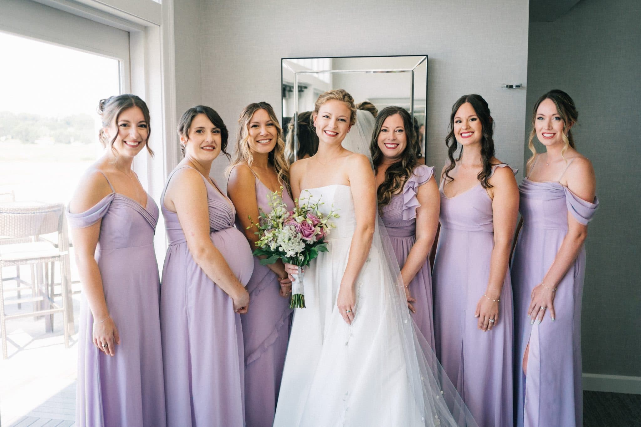 A bride in a white gown stands with six bridesmaids in lavender dresses, smiling in a well-lit room.
