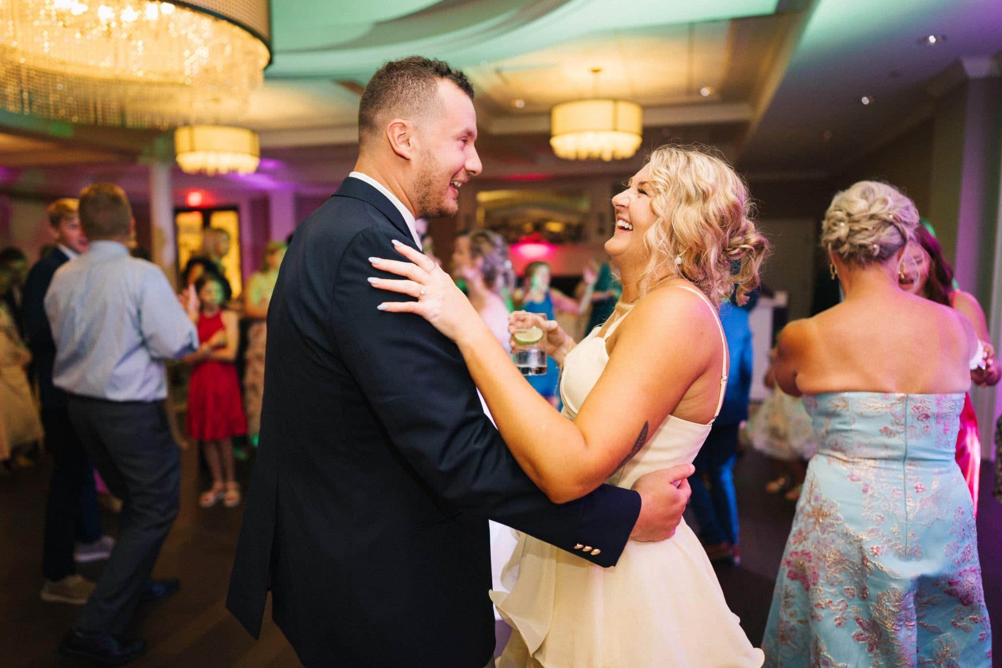 A couple dances closely in a brightly lit room with guests in the background.