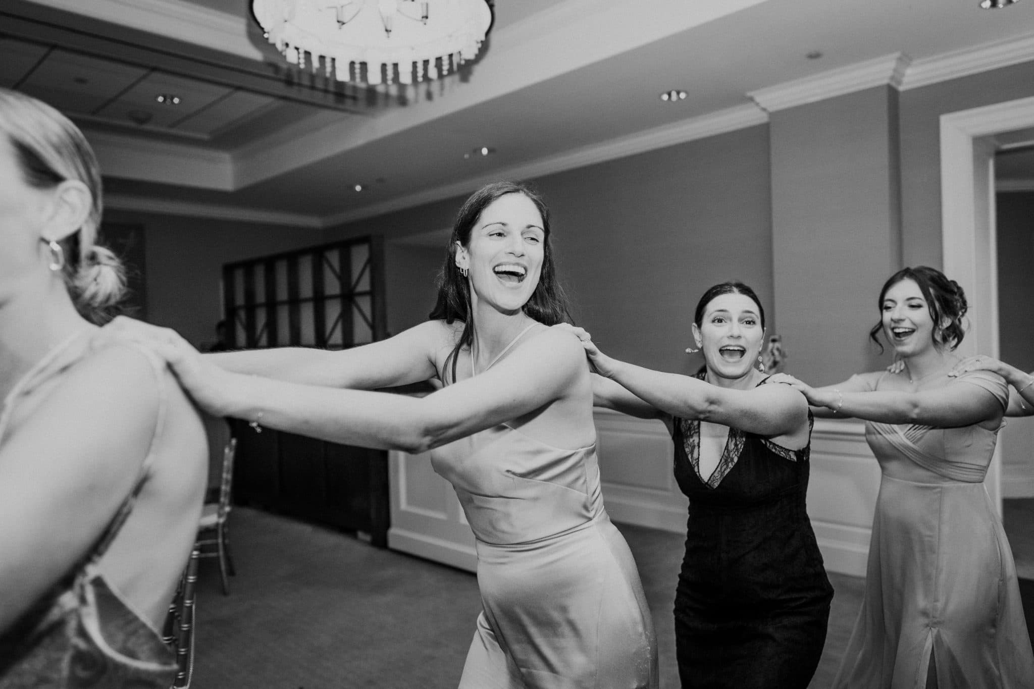 Four women in a room, one in a dress, others in formal attire, holding hands and smiling.