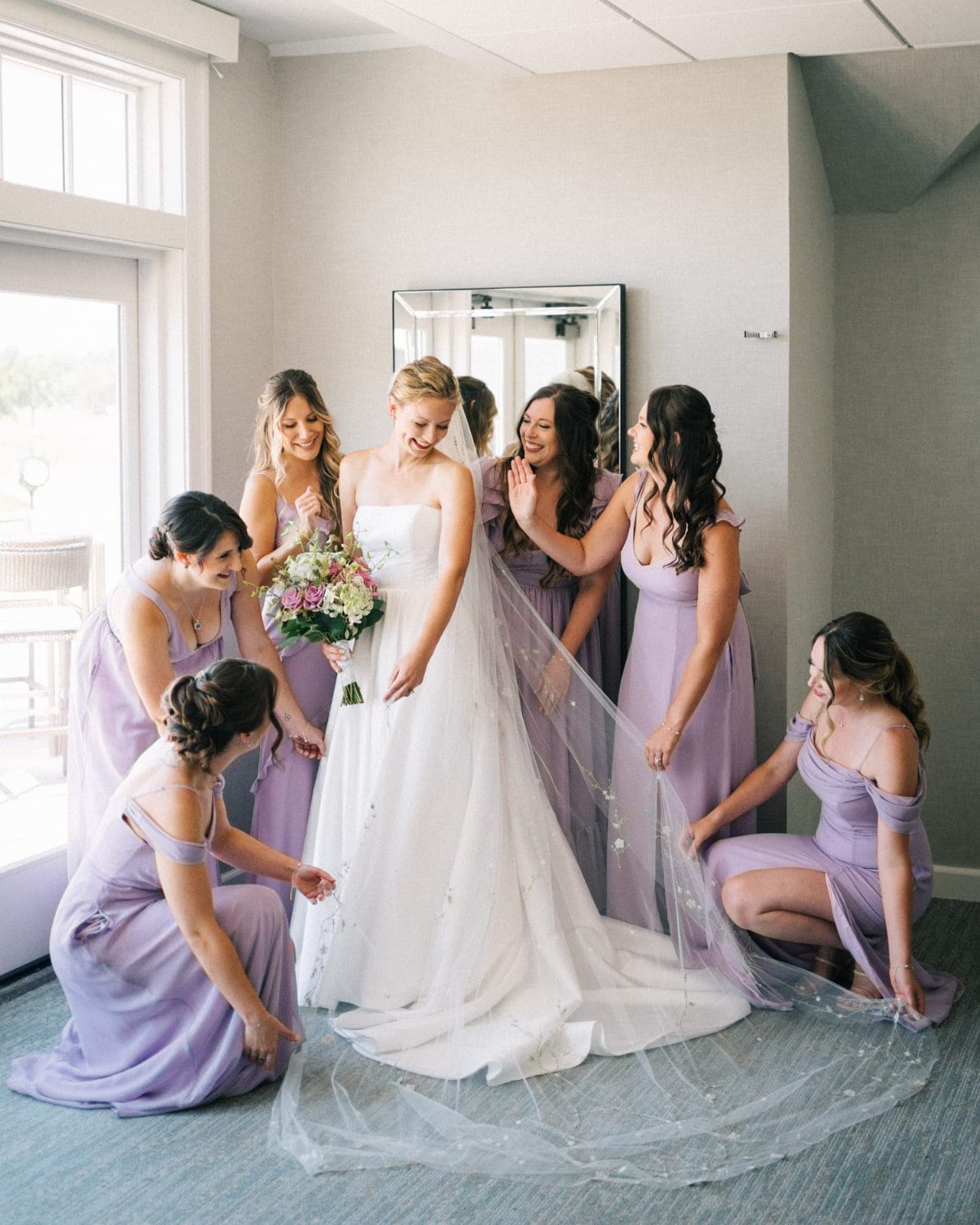 A bride in a white dress stands surrounded by bridesmaids in lavender dresses, adjusting her veil.